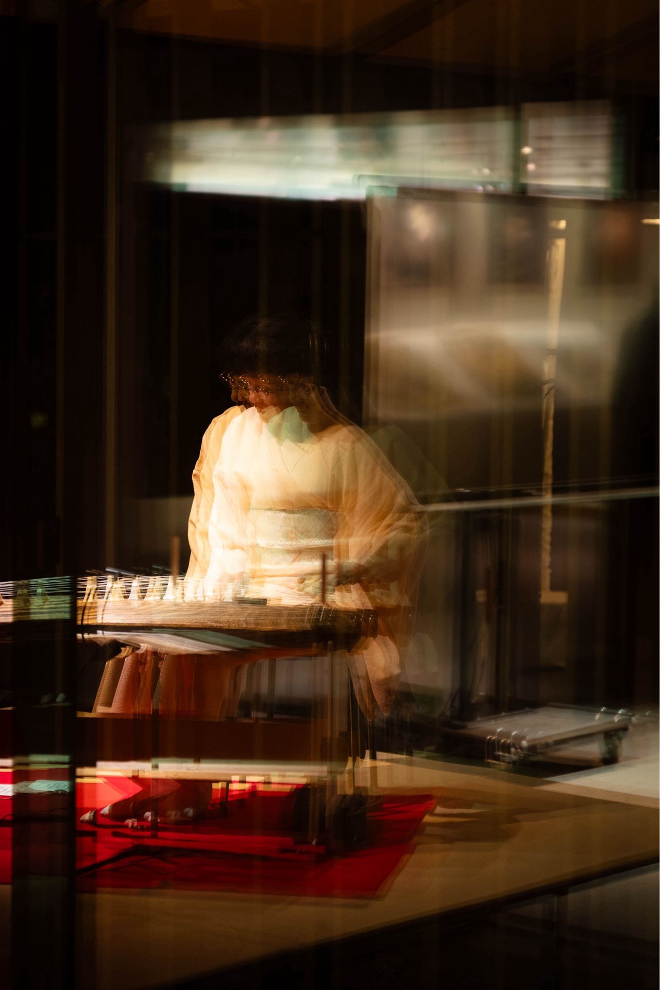 A blurred, ethereal view of a person in traditional Japanese attire playing a koto (a long Japanese zither) through a reflective surface. The image has a warm, golden glow, with the musician's form softly illuminated against a darker background.