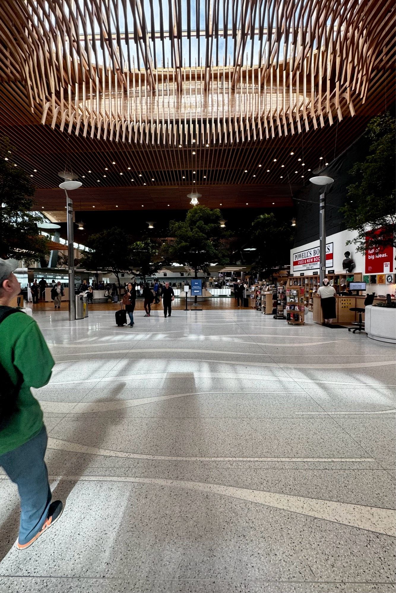 A wide view of an airport terminal interior showcasing a dramatic wooden ceiling with a central skylight. The space features a Powell's Books store, visible signage, and various retail areas. Travelers are walking through the open area with polished stone flooring. The design incorporates natural elements with indoor trees scattered throughout.