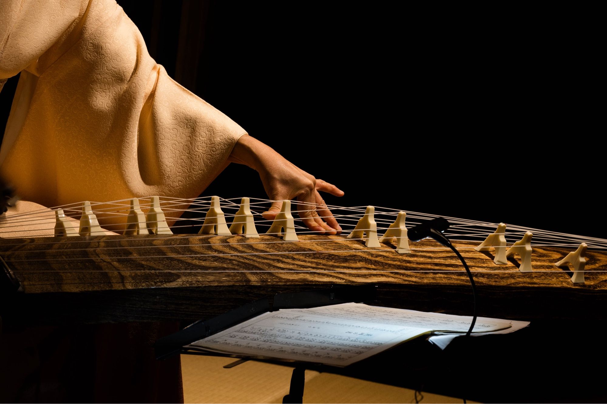 A close-up view of a person's hand and arm in a cream-colored kimono sleeve, plucking the strings of a koto. The wooden body of the instrument is visible, showing its intricate grain. A sheet of music is partially visible in the foreground.