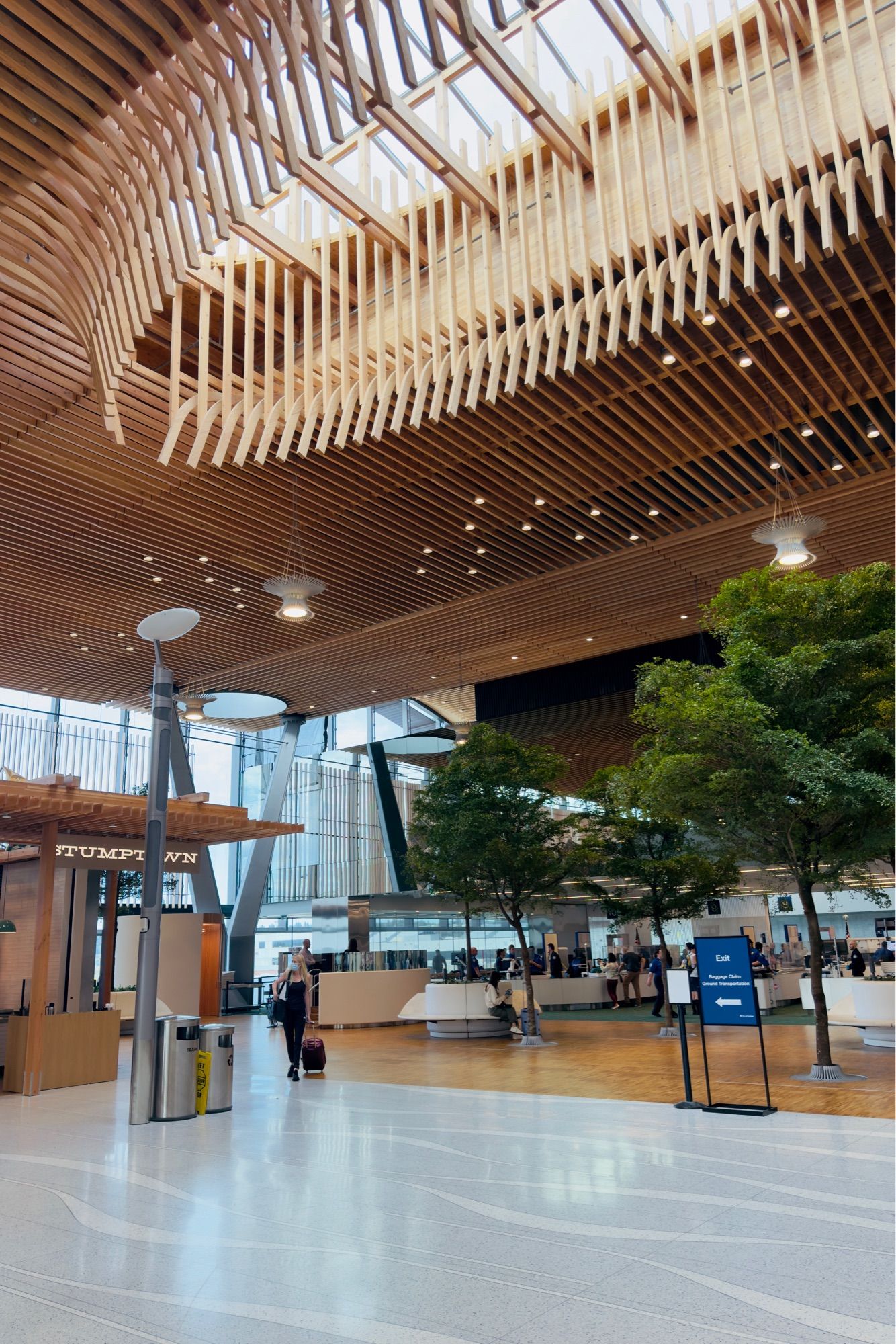 A modern airport terminal with an intricate wooden ceiling design featuring curved and latticed patterns. The space is well-lit with natural light from skylights and artificial lighting. Indoor trees and a Stumptown Coffee kiosk are visible. Passengers can be seen walking through the spacious area with reflective flooring.