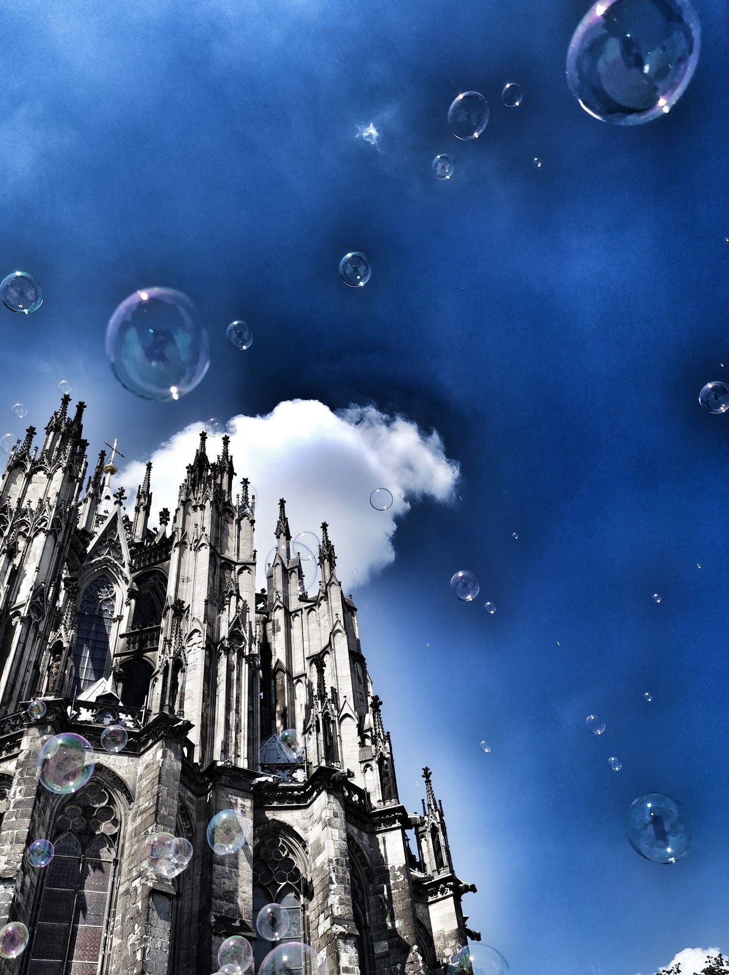 Teile des Kölner Doms vor einer weißen Wolke und blauem Himmel
Es fliegen Eeifenblasen herum und es sieht aus als würde es diese regnen