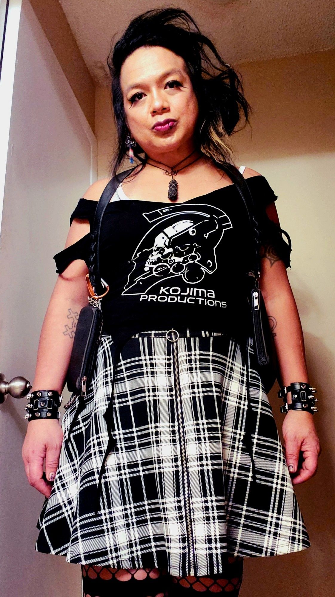 Kei standing shot from thighs up in banded black fishnets and a black and white plaid high waisted front zip skater skirt, black KOJIMA PRODUCTIONS LUDEN tee cut into a tendril hem crop with Bardot neck and chunk cut sleeve, black spiked wristbands and holster harness, black tourmaline and Estrogen chain, dreads back and hair teased but pinned and clipped on Her left, bathroom overhead lighting.