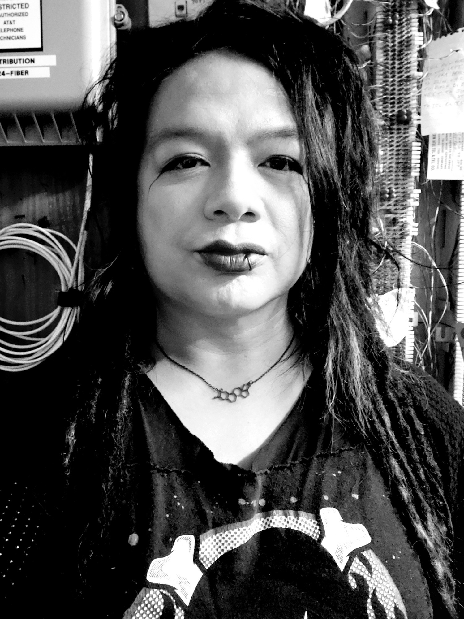 Kei shot from chest up in black cut YEAH YEAH YEAHS tee and textured shrug with Estrogen chain necklace, dark lipstick and thin extended puppy eyeliner, a few dreads forward on shoulders and hair loosely tamed, electrical panels behind Her and light for upper right of frame. 