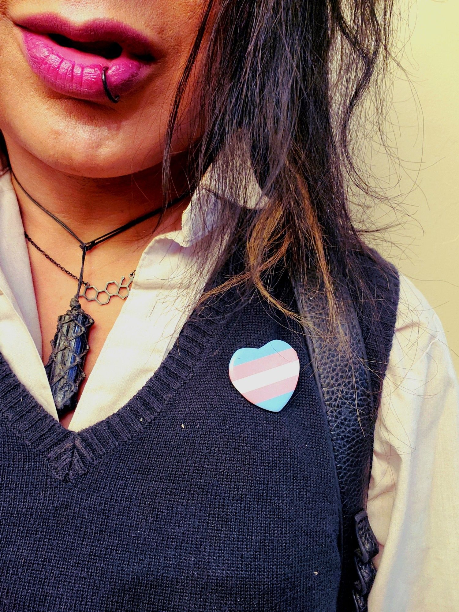 Closeup of Keinfrom chest to lips with Her roght shoulder out of frame, a Trans Pride pin in the shape of a heart in affixed to grey sweatervest over white button up, hair hanging into frame, black tourmaline and Estrogen chain with red lipstick.