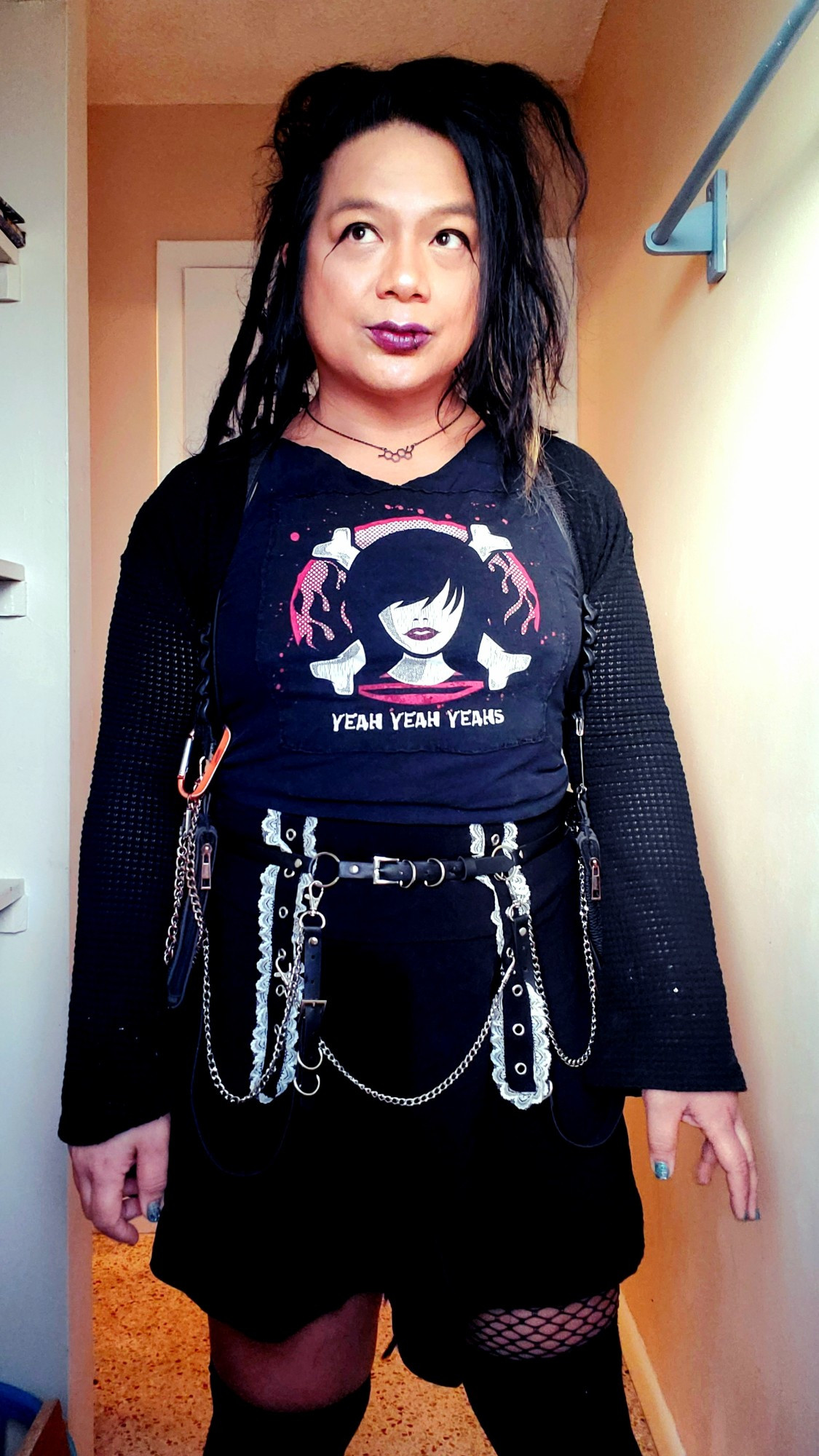 Kei standing shot from thighs up in black knit OverKneeSox with fishnet up left thigh, black skirt with front white lace straps and belt with chains and straps, cut YEAH YEAH YEAHS tee and a wide sleeve cuff textured black shrug, Estrogen chain and dark red lipstick with extended puppy eyeliner and a lotnof highlighter, dreads back and hair loosely tamed, backlit and bathroom window lighting.