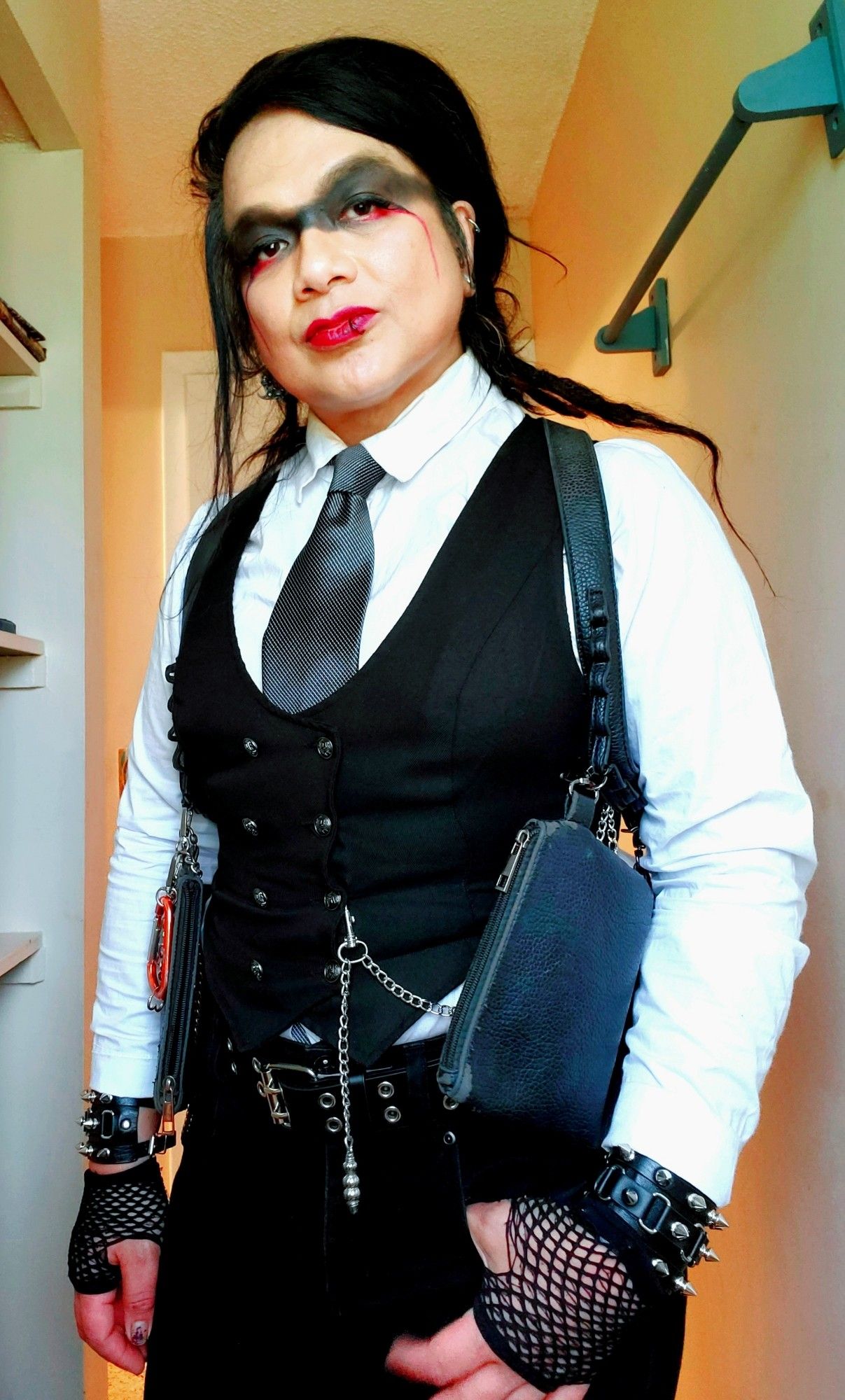 Kei from thighs up in black stinger jeans and double grommet belt, fingerless fishnet gloves and spiked bracelets, black double breastfed suit vest with chain, white LS button upnwith a gunmetal tie and holster harness, red lipstick and puppy eyeliner with red streaks down cheeks, dreads back and hair smoothed down right , bathroom window lighting.