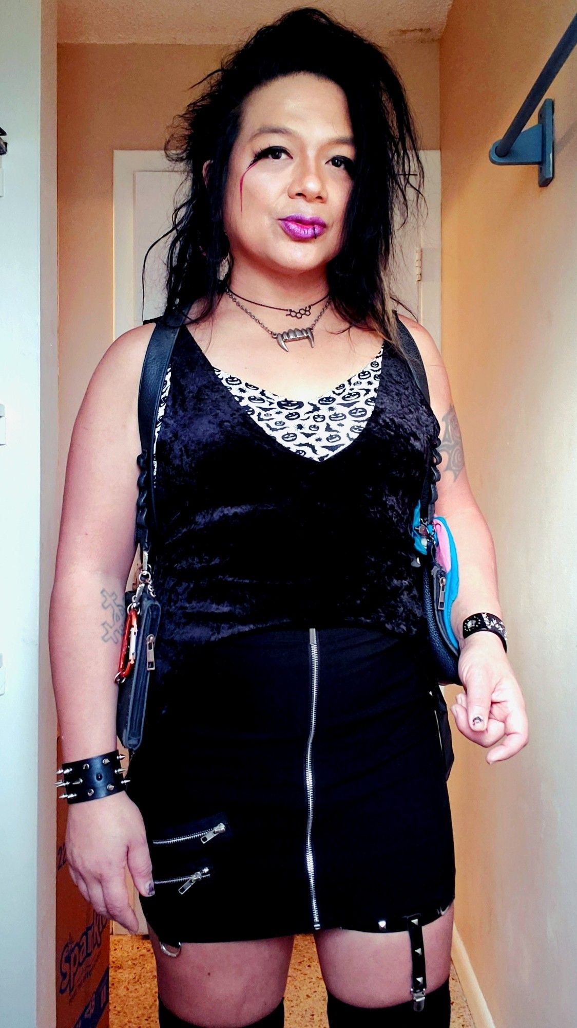 Kei standing shot from thighs up in black knit OverKneeSox with a studded garter on the left, black high-waisted front zip pencil skirt, jeweled WITCH and spiked cuffs, holster harness with Trans Pride Bandana tied on rig, black velvet top cut into a deep V cropped tank tied up in back, white with black Halliween print bra showing on chest and sides, chrome Fangs and Estrogen chain necklaces, dreads back and hair teased left, rednliostick with extra shimmer and puppy eyeliner with embellishments andbred streaks from outer corners and down cheeks, bathroom window lighting.