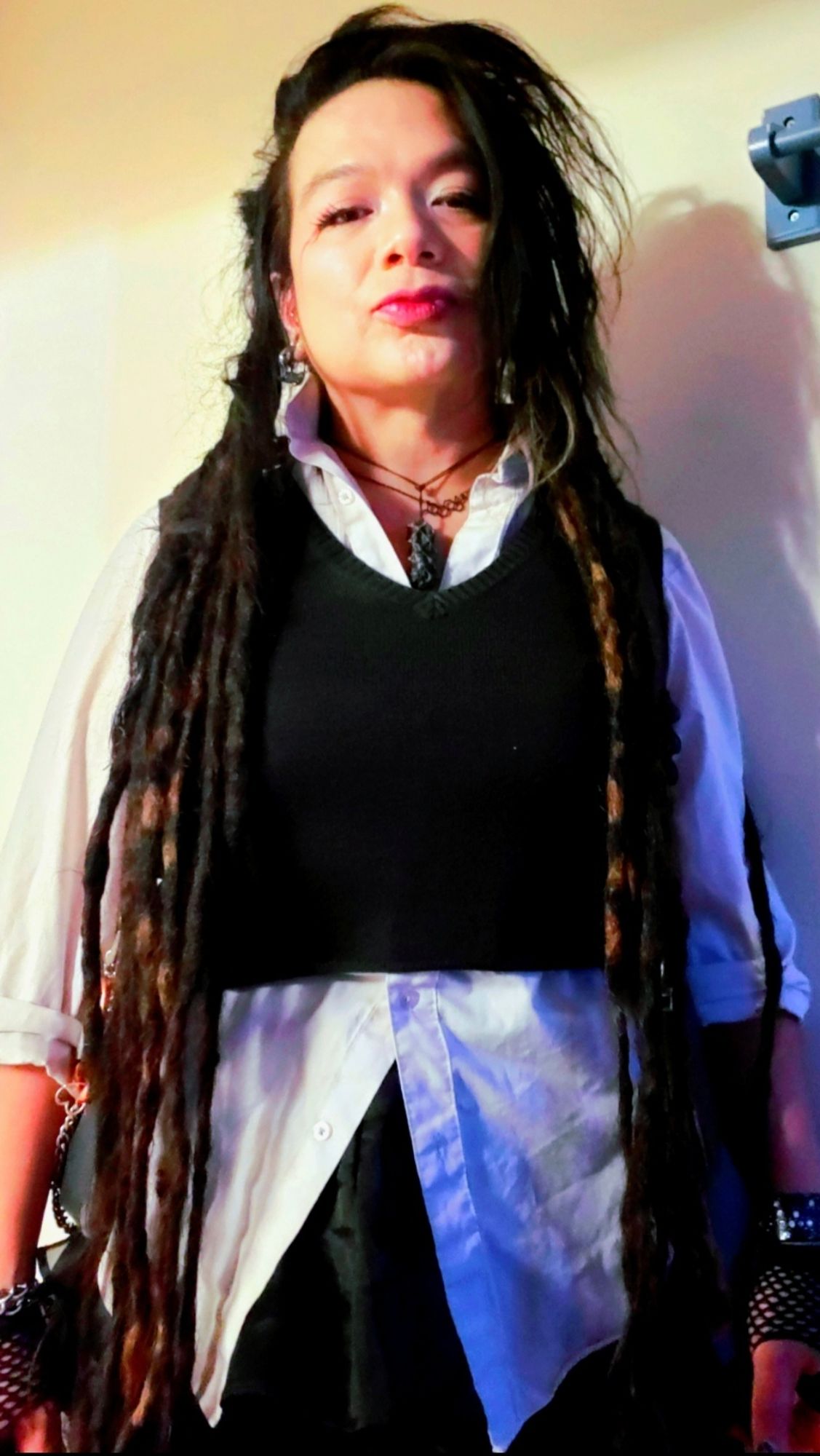 Kei from thighs up in black party skirt and white button up undone on top and bottom with sleeves flat rolled, fishnet fingerless gloves and jeweled WITCH cuff, charcoal sweatervest crop cut, black tourmaline and Estrogen chain, dreads forward on shoulders and hair flipped left, red kiostick and puppy eyeliner, bathroom side lighting.