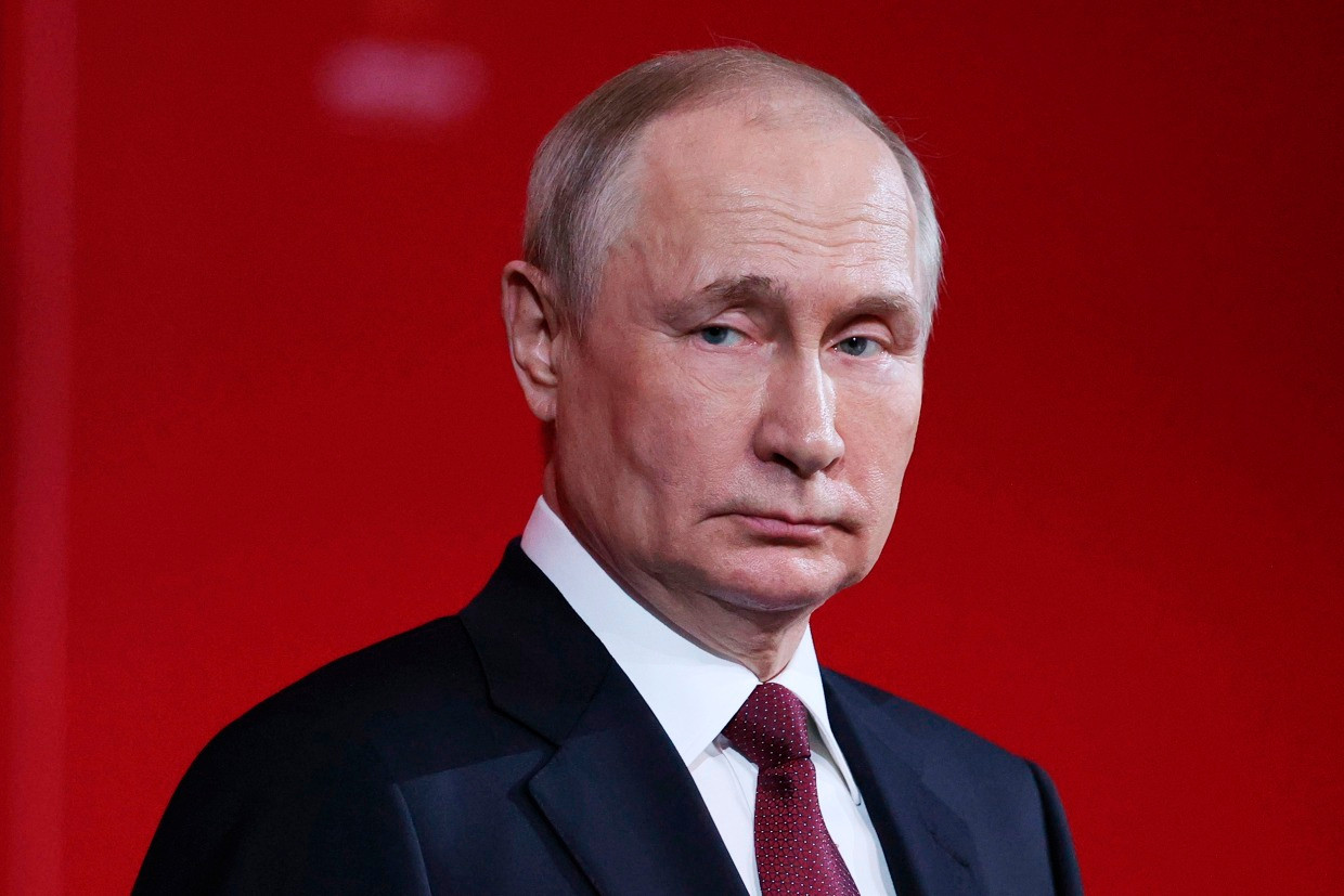 Vladimir Putin, looking serious in a suit, against a red background.