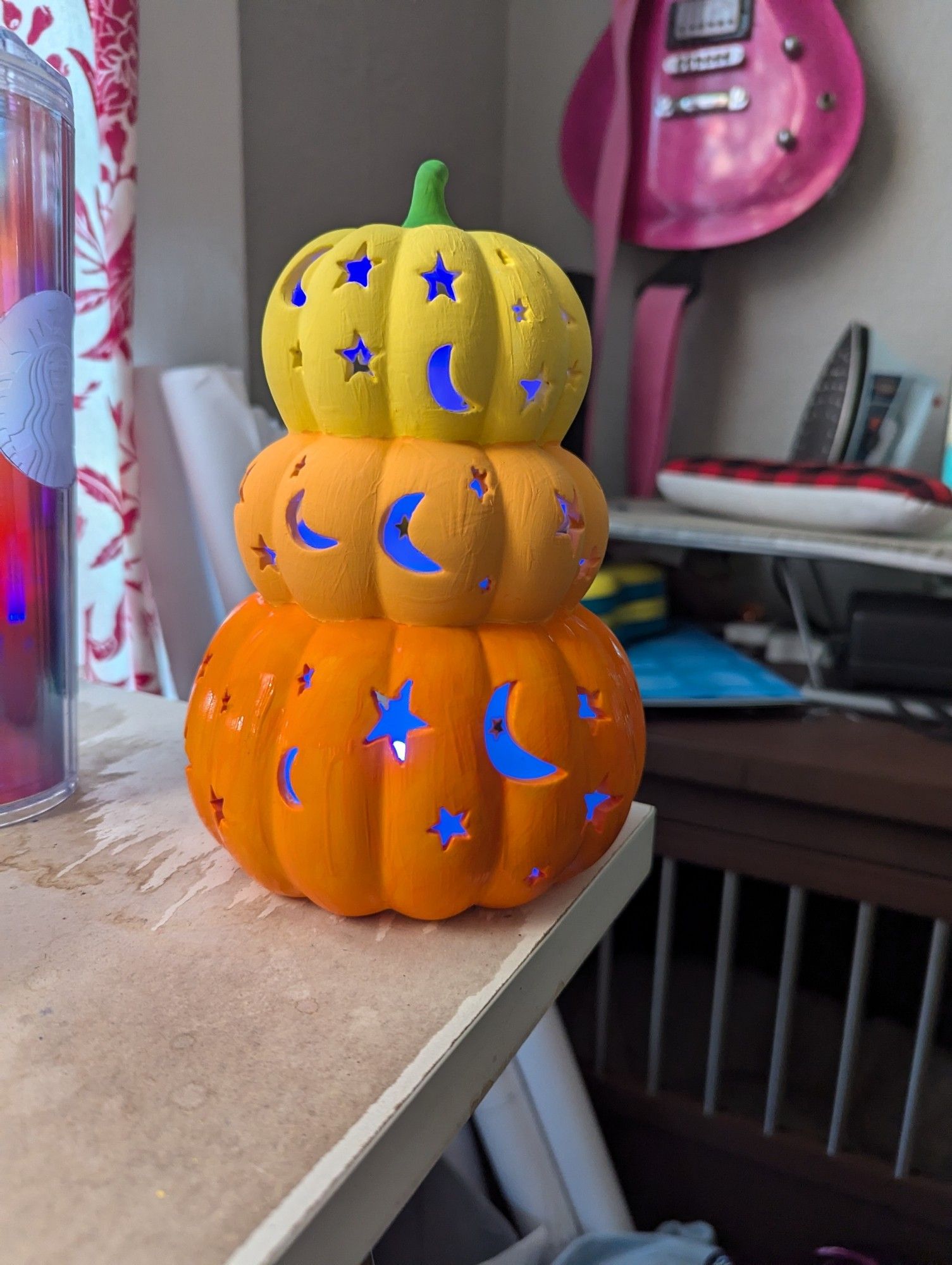 The same ceramic statue of three pumpkins stacked on top of each other, of descending size, painted yellow, light orange, and orange, now with a blue light illuminating the cutouts.