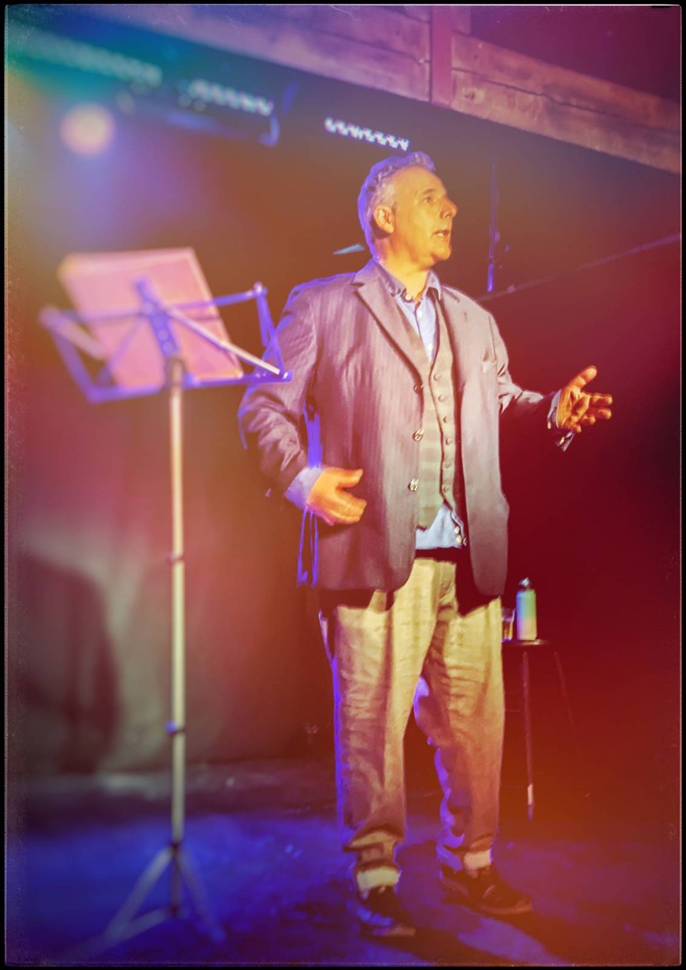 Rob Newman, in a shirt waistcoat and jacket gestures in front of a music stand full of notes.