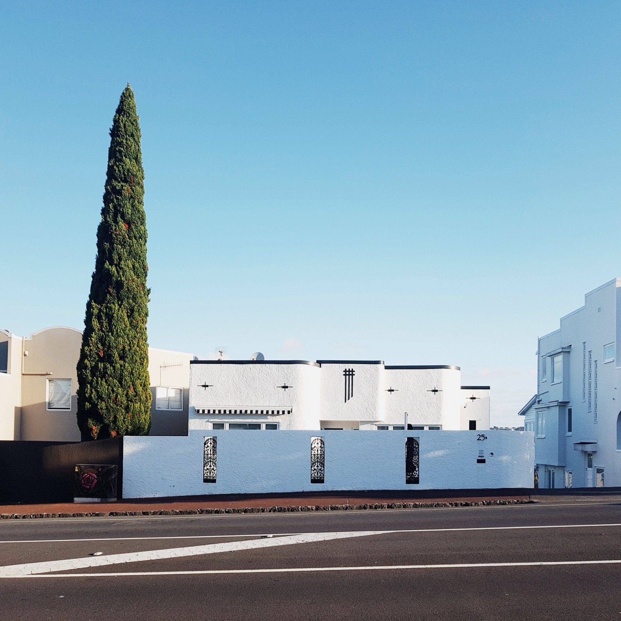 Architectural photography: a bright view of horizontal white mediterranean house. Cypress tree as a strong vertical element. A Road in the foreground, blue sky in the background 