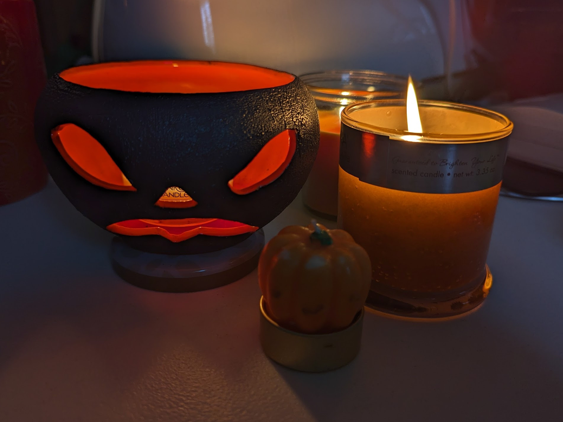 A small candle shrine featuring: a black ceramic jack o'lantern with a grimacing face and orange light spilling out, 2 orange candles in glass jars, and a small wax pumpkin candle (unlit)