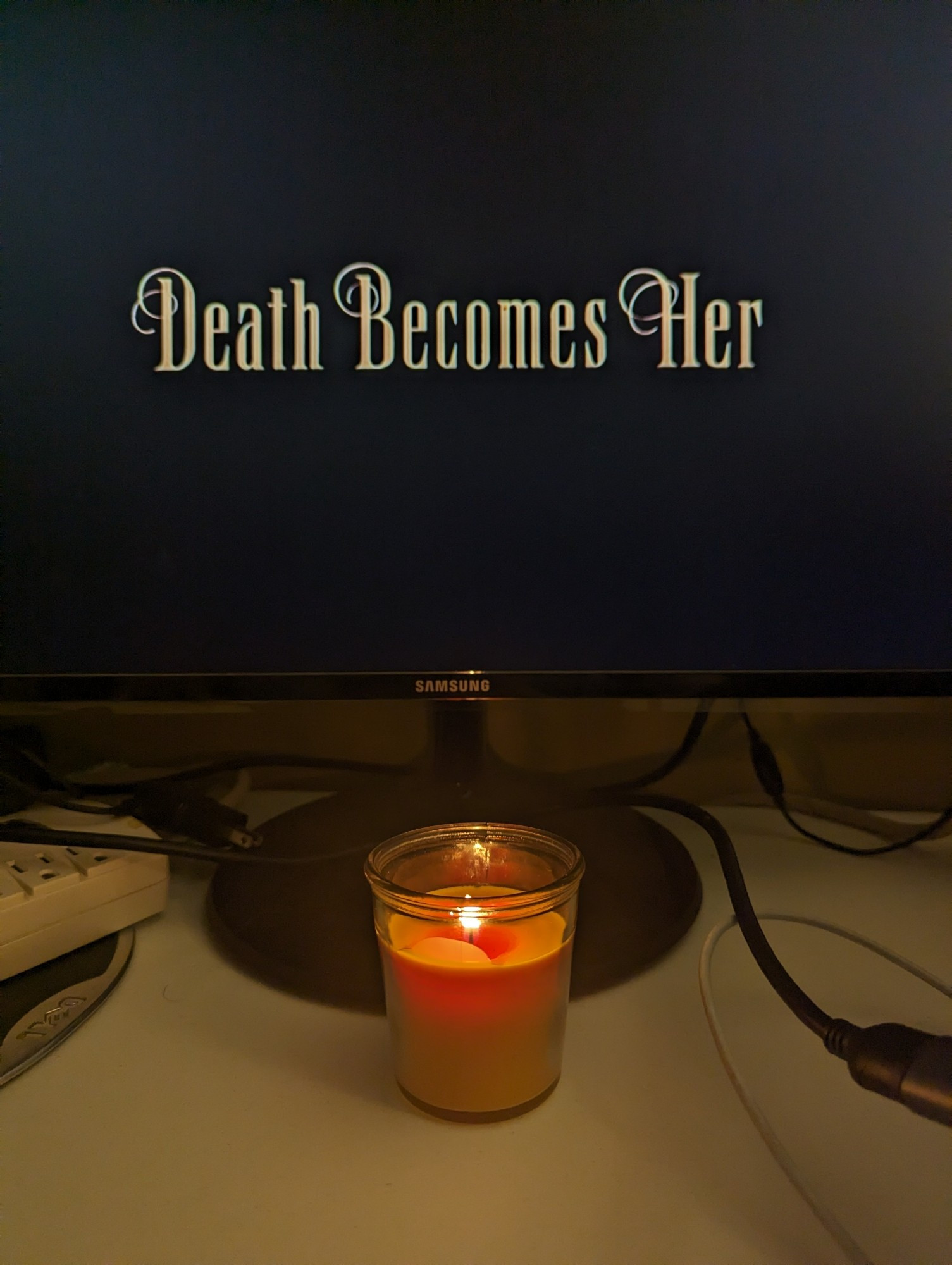 An orange candle below a monitor. On the monitor the text reads Death Becomes Her