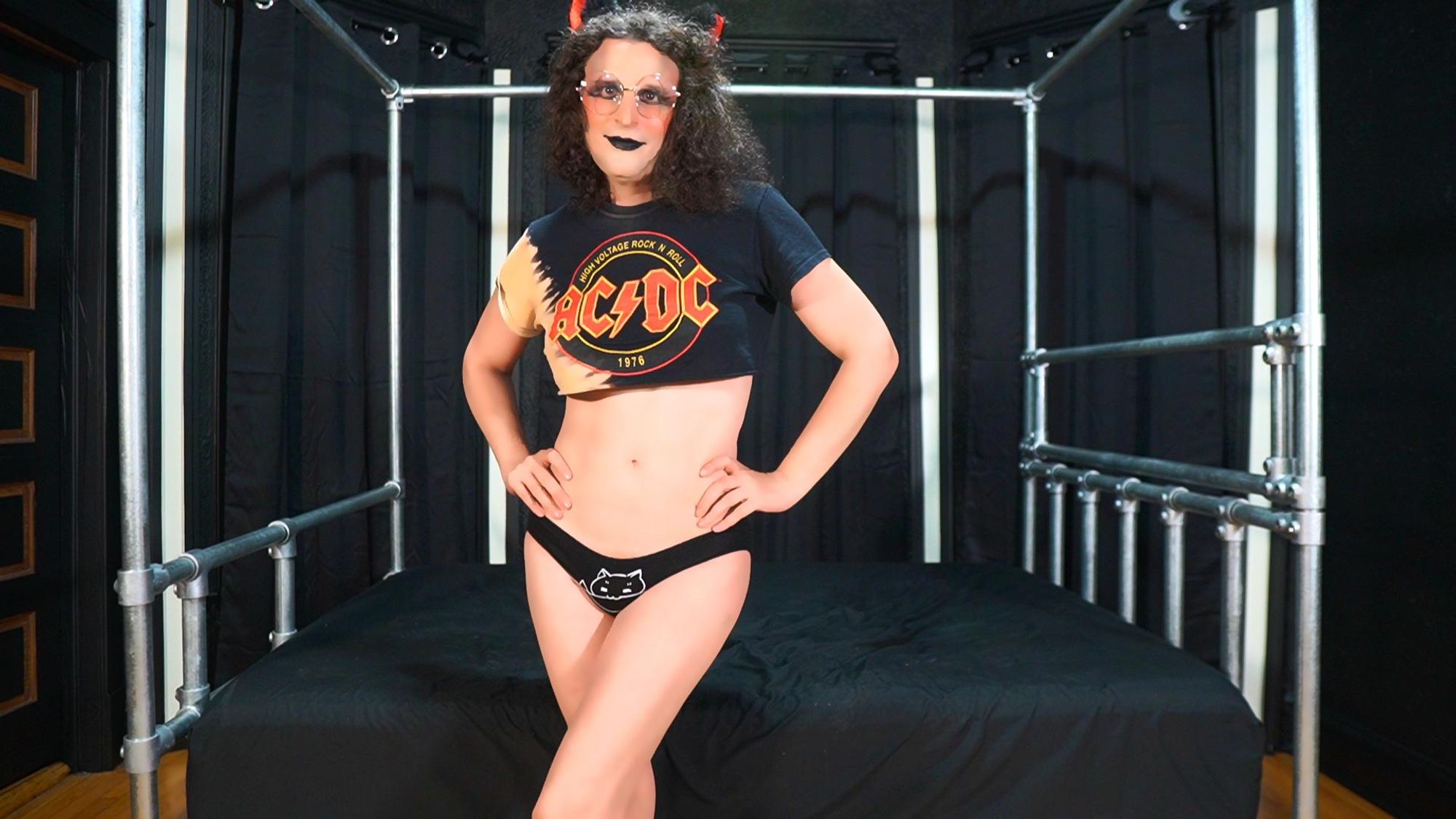 A woman with long brown hair is posing for a picture in a bedroom. She is wearing a black shirt with a red and yellow AC/DC logo on it, as well as black panties. The room has a bed with black sheets and a large window with a large black curtain. The woman is standing in front of the window, with her hands on her hips and a confident expression on her face. The overall color scheme of the room is dark with shades of black, gray, and brown. The woman appears to be in her thirties and is exuding a sense of empowerment and independence.