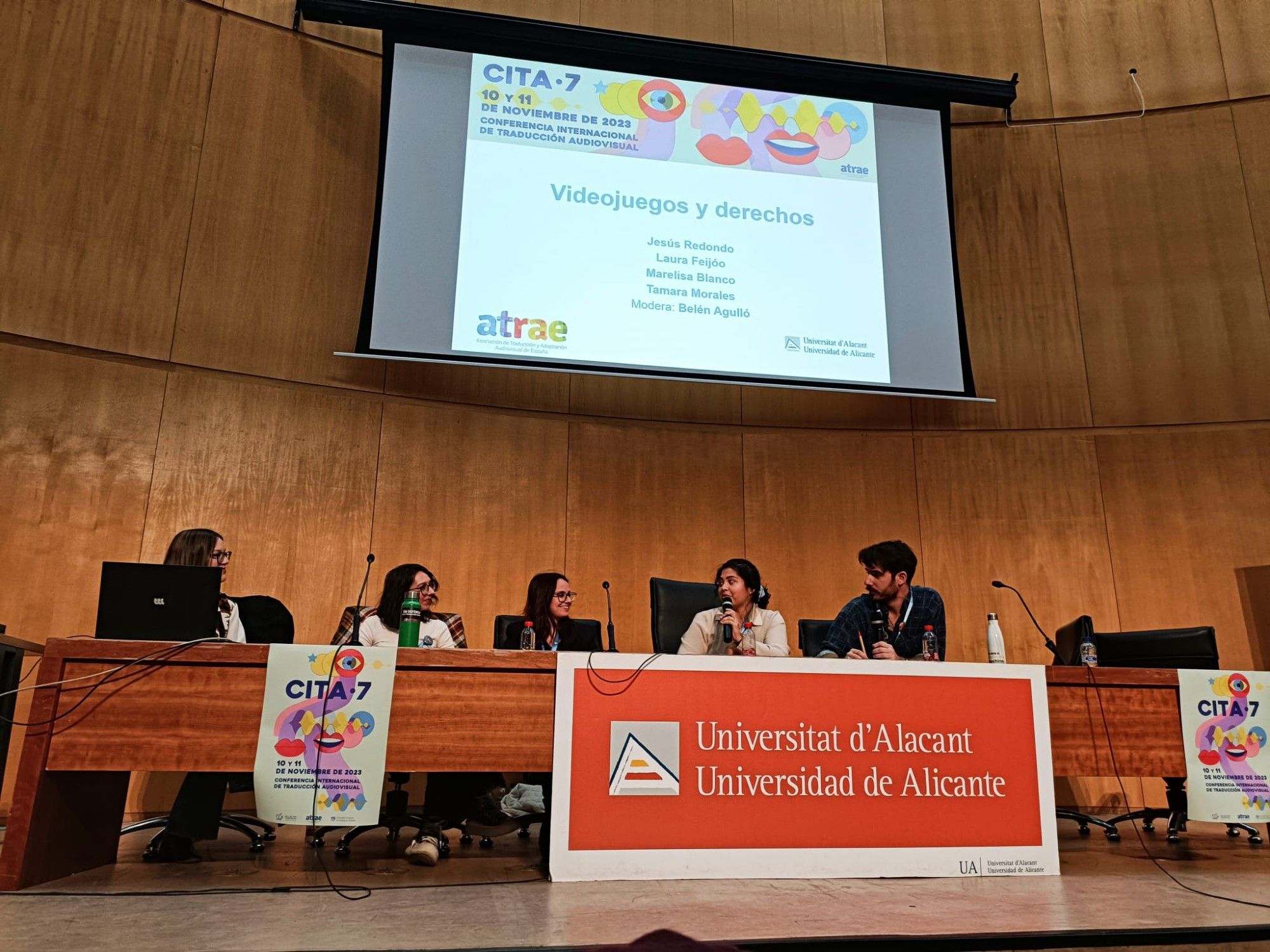 Sentades en una mesa que contiene carteles con los logos de la Universidad de Alicante-Universitat d'Alacant, la CITA, la asociación ATRAE de DAMA Autor, hay cinco personas. De izquierda a derecha, Belén Agulló (moderadora de la mesa), Tamara Morales (traductora autónoma, miembro de la junta y vocal de la Comisión de Videojuegos), Laura Feijóo (traductora autónoma, miembro de la Comisión de Premios), Marelisa Blanco (abogada especialista en derechos de autor y propiedad intelectual) y Jesús Redondo (gestor de proyectos de localización de videojuegos en Native Prime).
En la parte superior de la sala, hay una pantalla que muestra el título de la mesa redonda, los nombres de les ponentes, el logo de ATRAE y el de la Universidad de Alicante, y también una imagen que incluye parte del cartel de la CITA 7, que se basa en dos bocas, una abierta y otra cerrada, un ojo con un iris de color azul, y una onda de sonido.