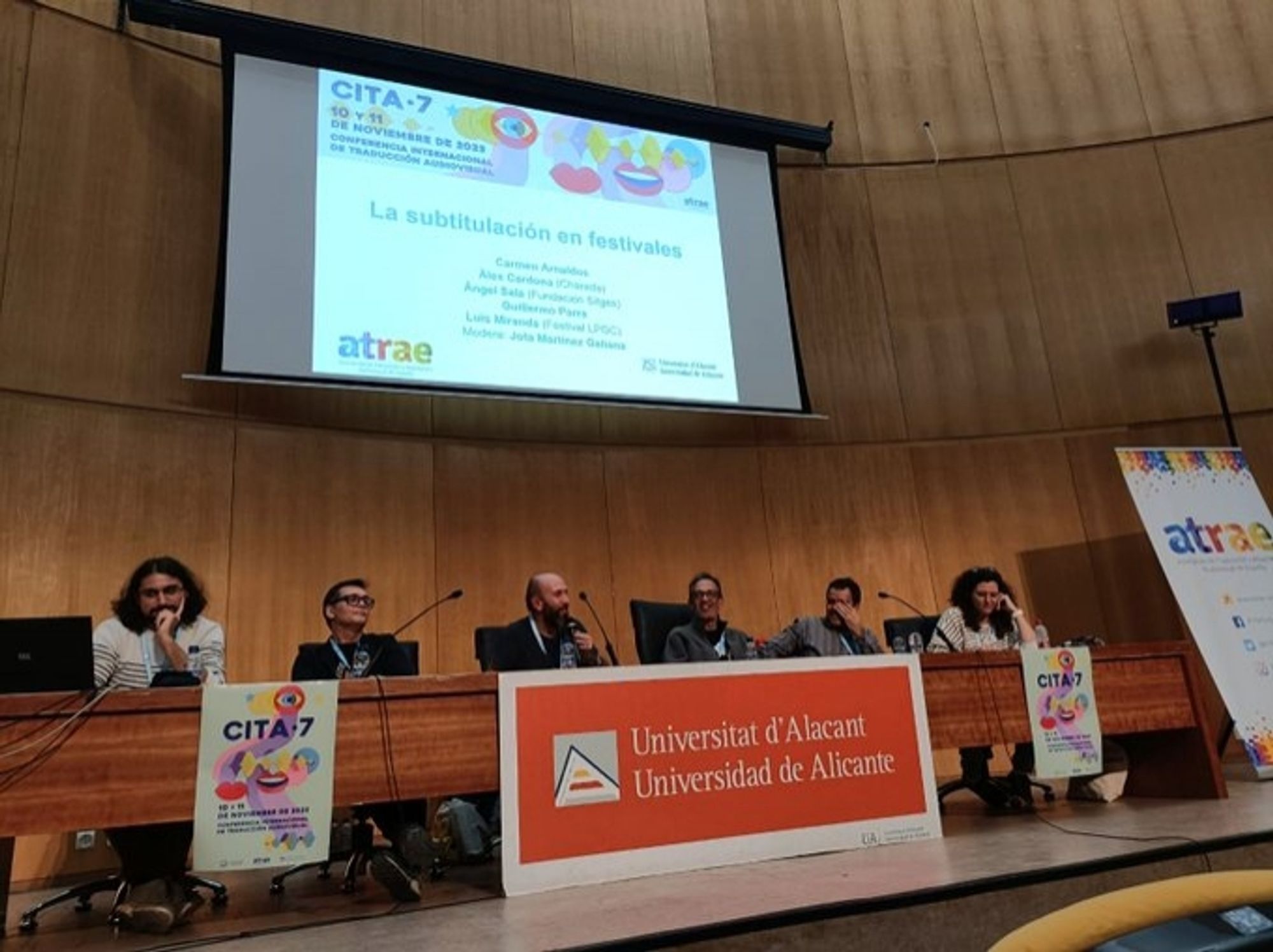 Sentades en una mesa que contiene carteles con los logos de la Universidad de Alicante-Universitat d'Alacant, la CITA, la asociación ATRAE y de DAMA Autor, hay cincos hombres y una mujer. De izquierda a derecha. Guillermo Parra (traductor autónomo coordinador del Festival
Americana), Ángel Sala (director del Festival Sitges), Jota Martínez
Galiana (moderador de la mesa), Luis Miranda (director del Festival Internacional de Cine de Las Palmas de Gran Canaria), Àlex Cardona (socio fundador de Charada, empresa dedicada a la traducción audiovisual, y se encarga de la supervisión de proyectos) y Carmen Arnaldos, traductora audiovisual autónoma. En la parte superior de la sala, hay una pantalla que muestra el título de la mesa redonda, los nombres de les ponentes, el logo de ATRAE y el de la Universidad de Alicante, y también una imagen que incluye parte del cartel de la CITA 7, que se basa en dos bocas, una cerrada y otra abierta, un ojo con un iris de color azul, y una onda de sonido.