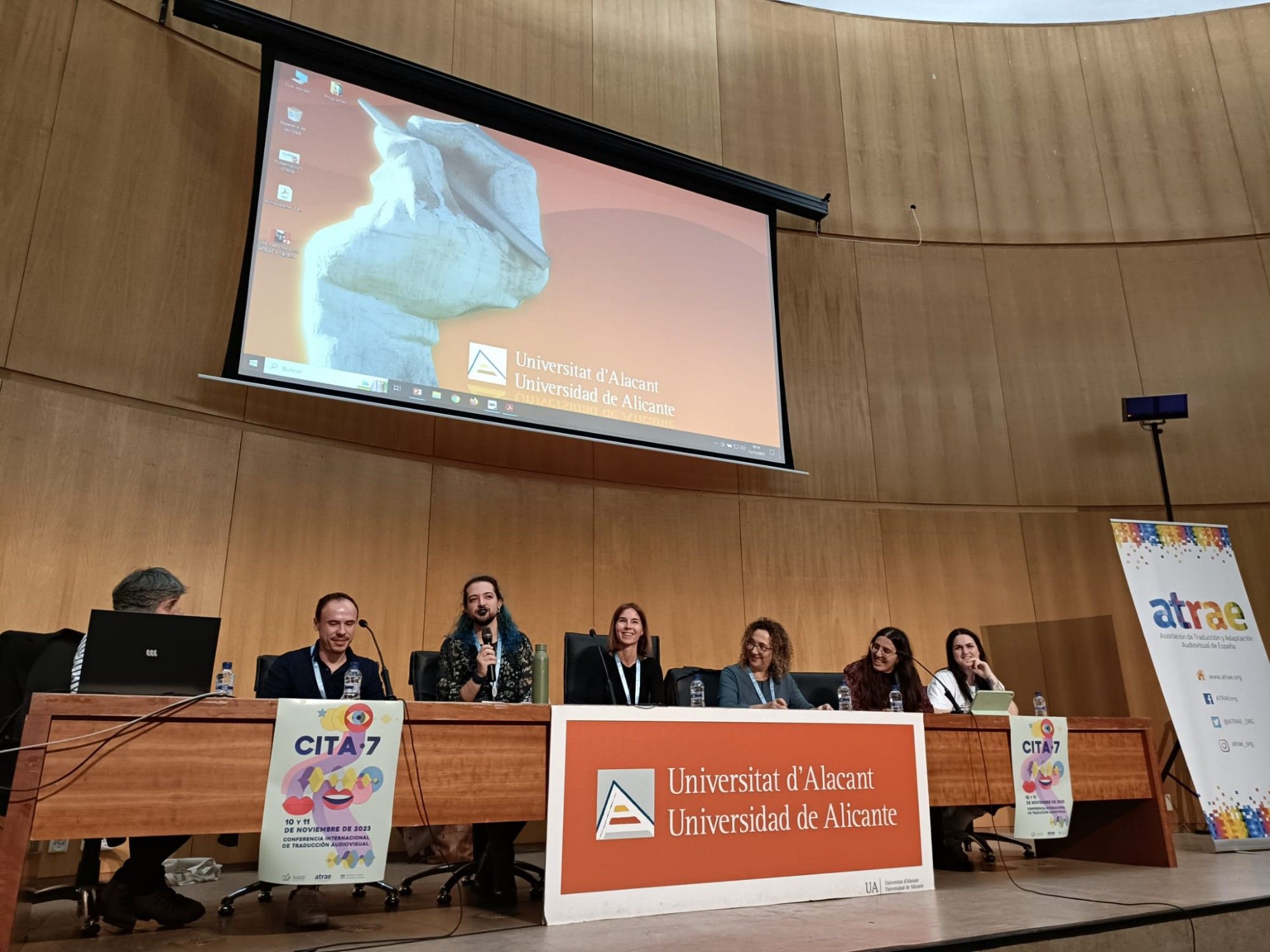 Sentades en una mesa que contiene carteles con los logos de la Universidad de Alicante-Universitat d'Alacant, la CITA, la asociación ATRAE y de DAMA Autor, hay cinco ponentes. De izquierda a derecha, Beatriz Zabalondo, Antonio Martínez Pleguezuelos, Ártemis López, Carla Botella (que modera la mesa), Eva Espasa, Irene Vilchez y Carla Míguez.
En la parte superior de la sala, hay una pantalla que muestra la escultura que se encuentra en la entrada de la universidad. Dicha escultura es una mano gigante que coge un lápiz.