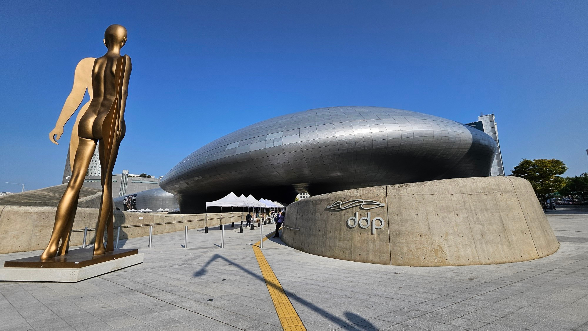 The futuristic silver building in the area affectionately called the DDP (the Dongdaemun Design Plaza) with a extremely tall bronze colored statue to the left 