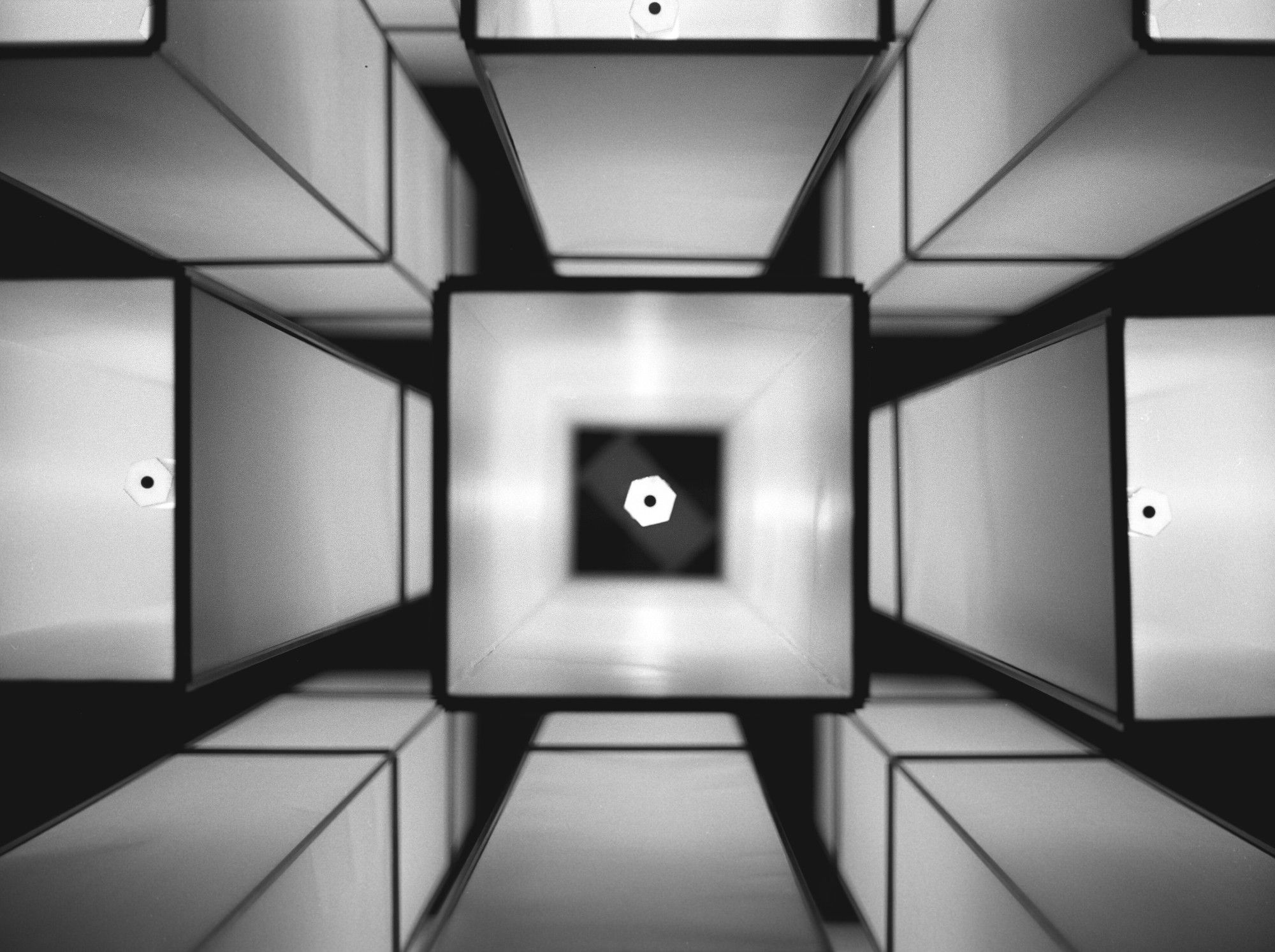 A photo looking upwards at the bottom of a bunch of long, tall light fixtures, centering on the element in the middle of one of the square tubes. It's black and white. It's a repeating pattern with a vanishing point