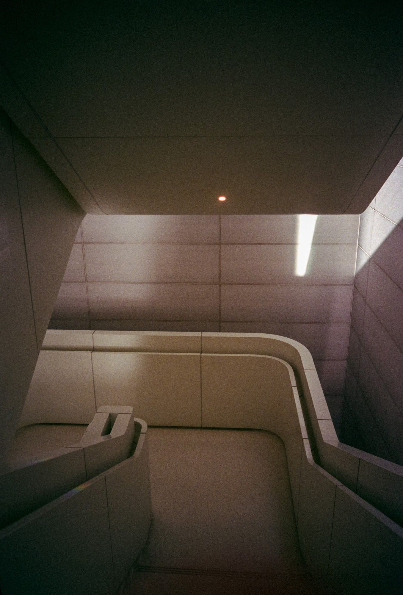 A photo of a clean and dimly lit modernish brutalist staircase with clean curves at the corner of the hand rails, but otherwise entirely straight panels for the ceilings, walls, handrails, and floor.