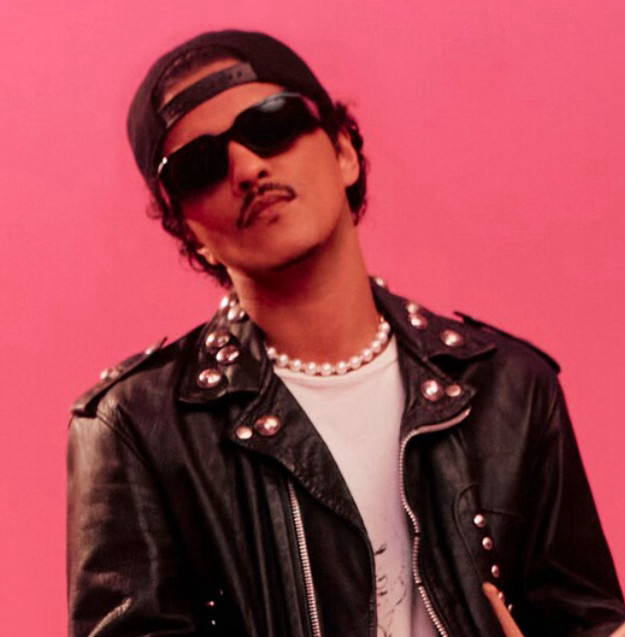 Bruno Mars wearing a black backwards baseball cap and black sunglasses, with a white t-shirt and black leather jacket covered in studs and zips a la ‘Bad’ era Michael Jackson. He has a moustache highly reminiscent of the one Weird Al sported at the time he parodied ‘Bad’. For some reason he is also wearing a chunky white pearl necklace like you’d get free in a cracker and wear when you were a child too young to know better. All that money and that’s what he picks.