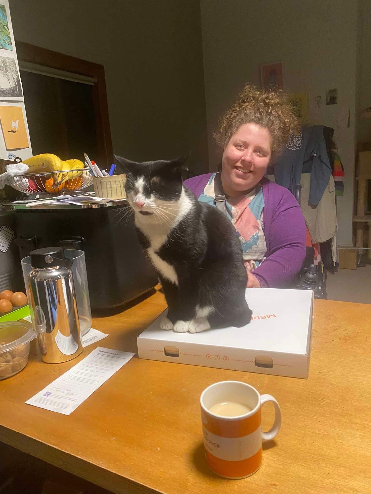 A black and white cat sitting on top of a pizza box with me smiling in the background.