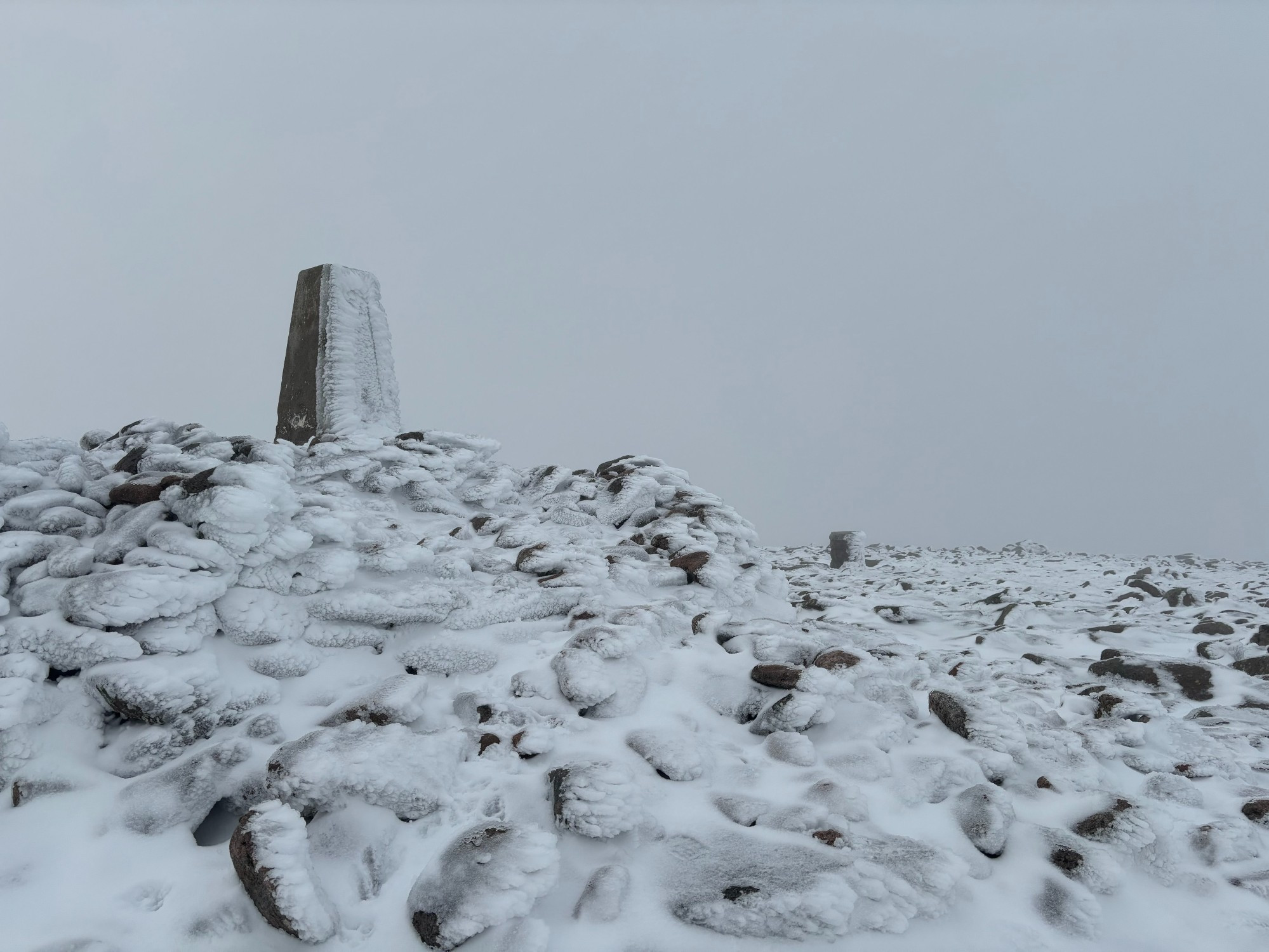 The trig point and summit cairn of Ben Macdui covered in snow.