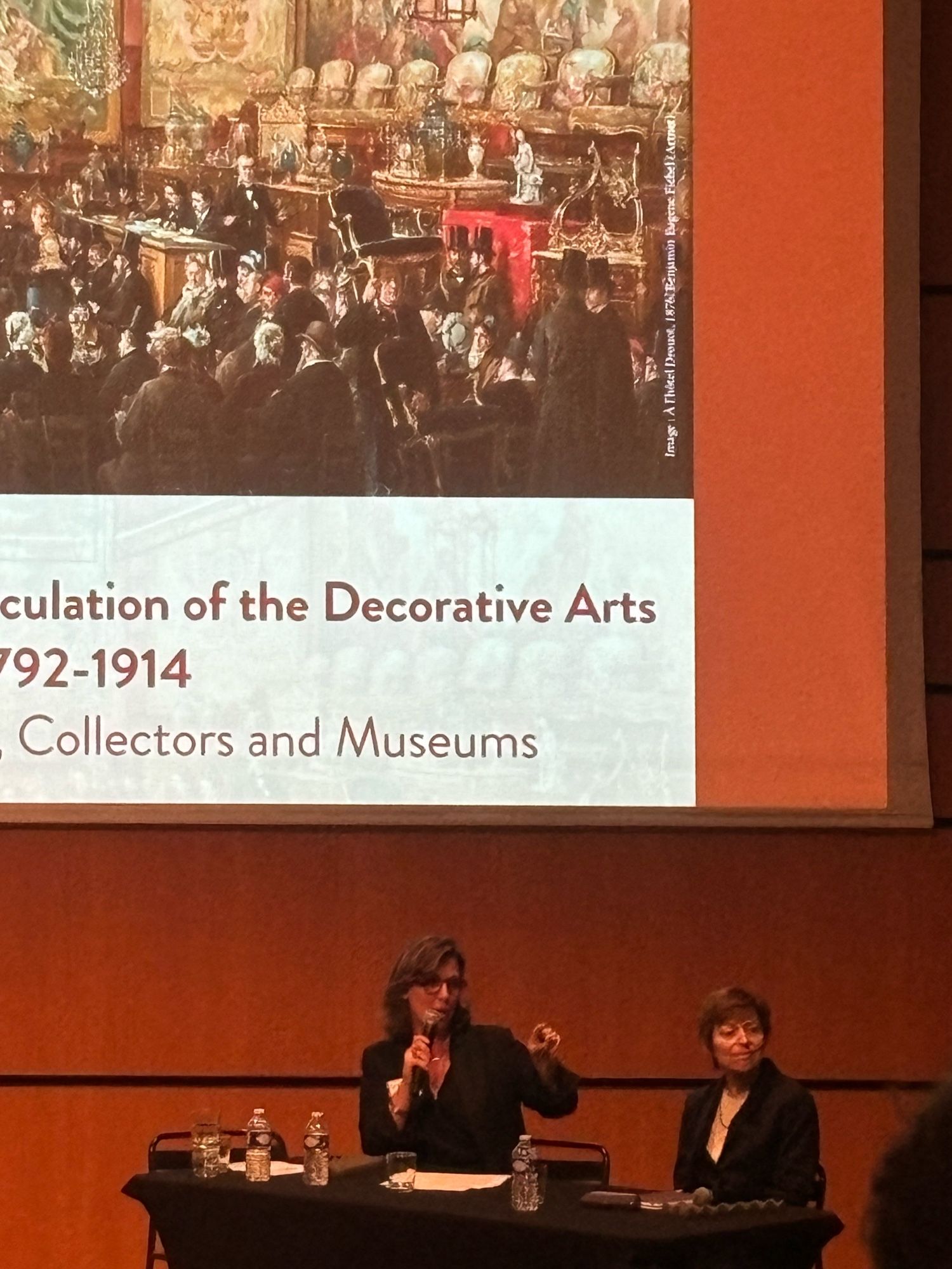 Camille Mestdah on the left and Diana Davies on the right, talking below the powerpoint displaying the conference poster, an image of a nineteenth century auction in Drouot - bathed in lush red light, taken from the comfort of the theatre like auditorium