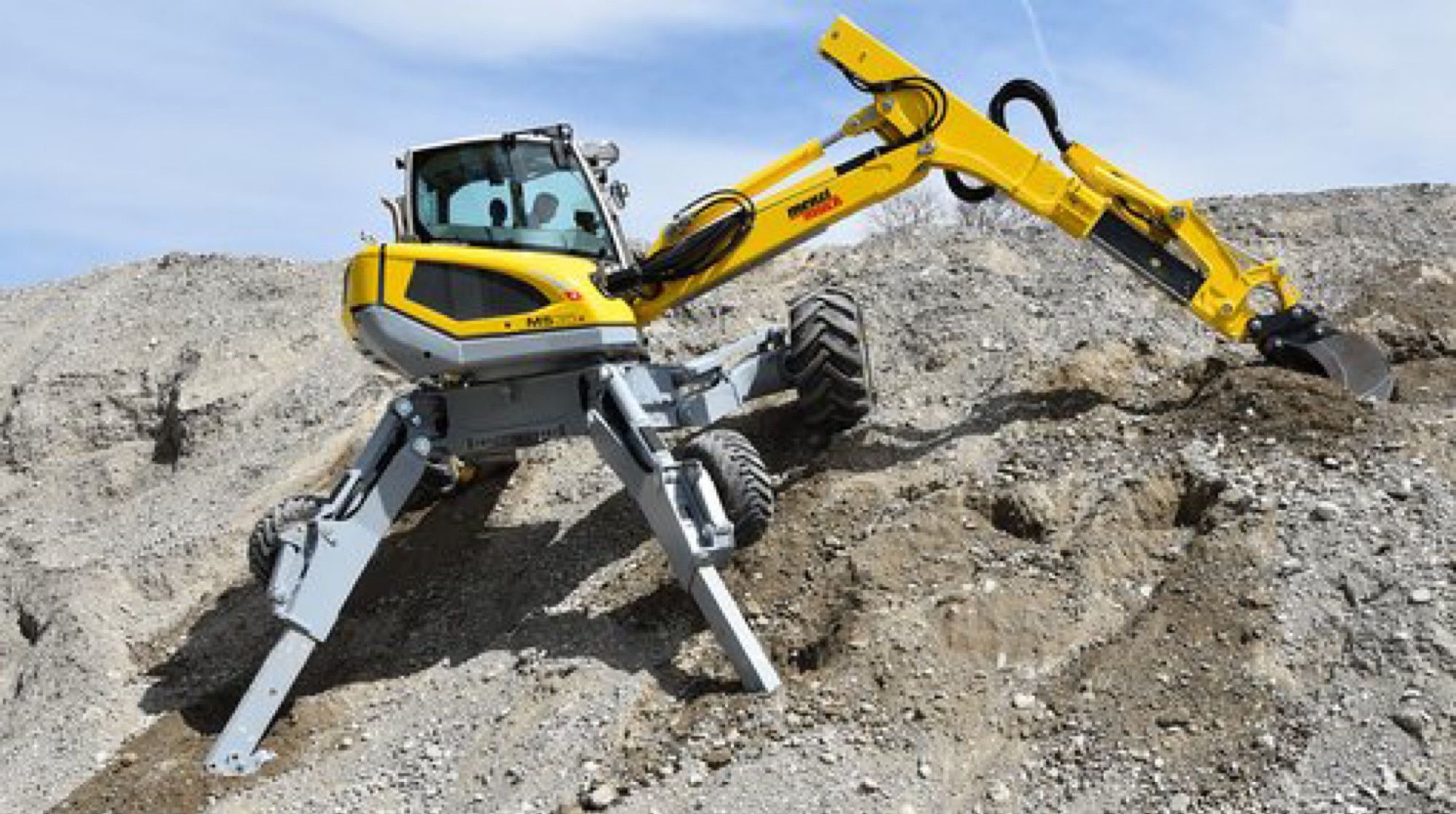 A spider excavator with its legs extended. It is scooping rubble from a giant rock pile