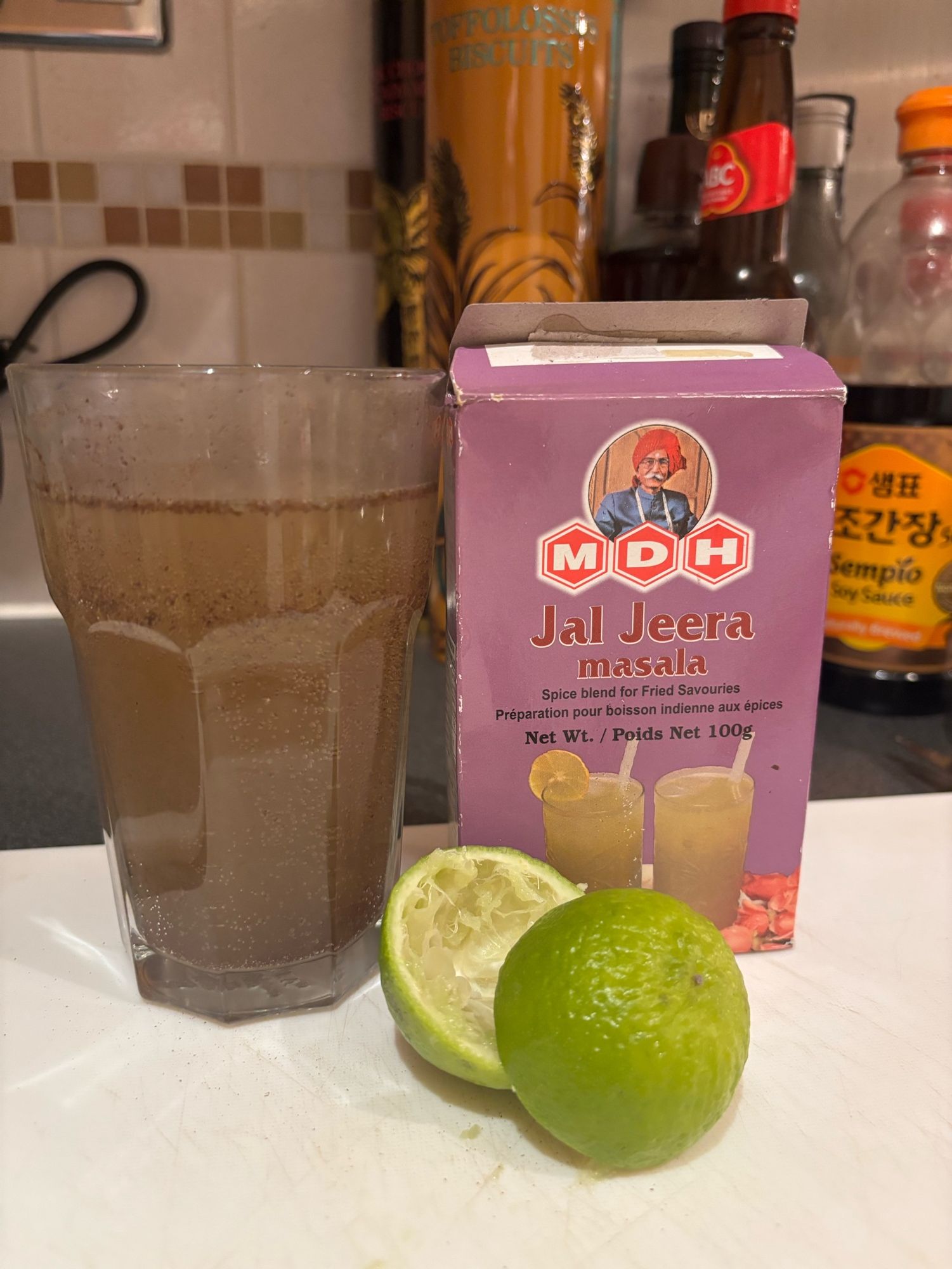 A glass filled with a brownish liquid, next to it a purple box labelled “Jal Jeera” and two halves of a squeezed lime 