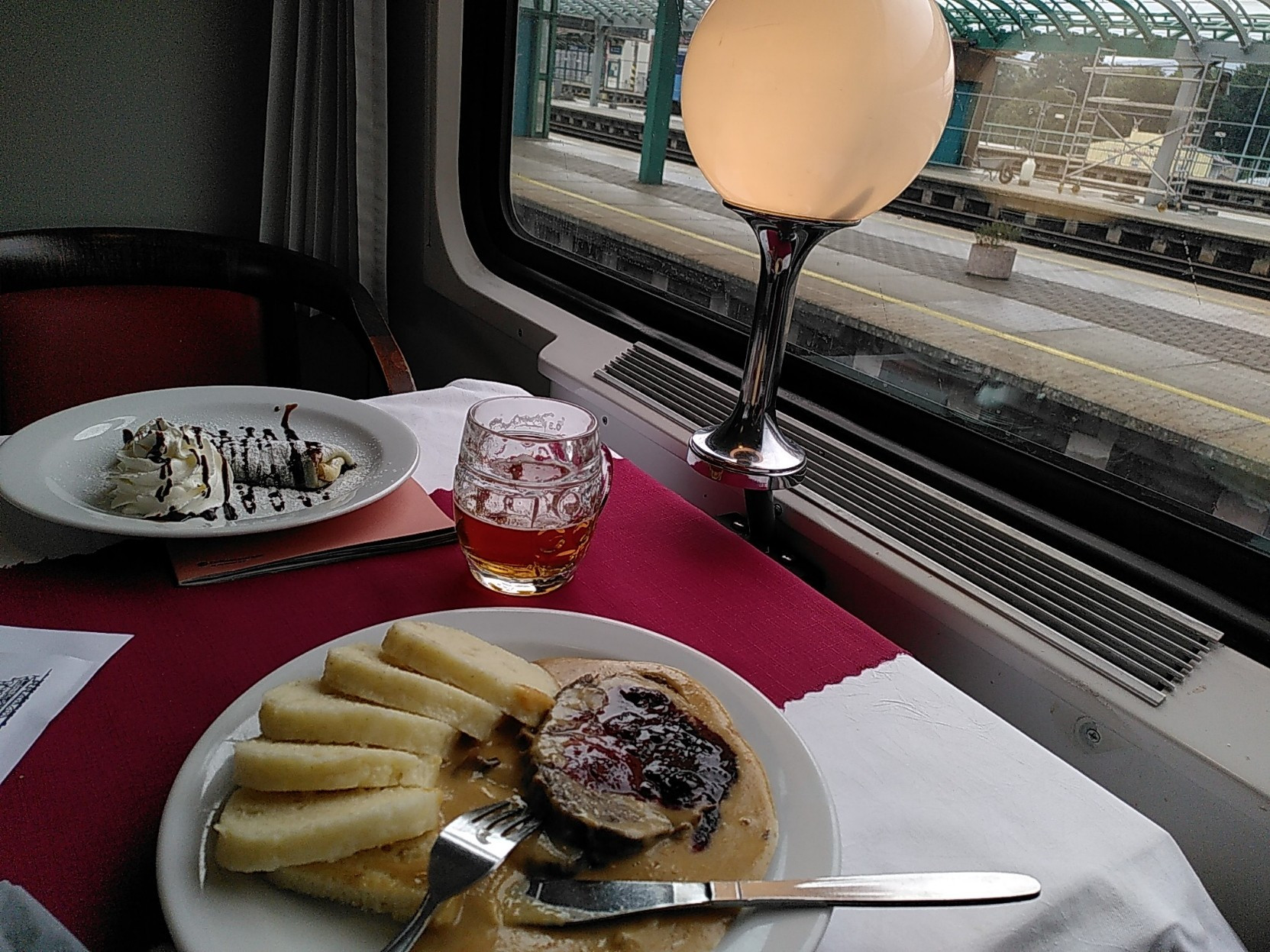 Blick auf Tisch im Zugrestaurant, am vorderen Teller Lendenbraten, am hinteren Schokoladepalatschinke, dazwischen Bier.
Vor dem Fenster befindet sich am Tisch montiert eine kleine Leuchte, draussen sind Bahnsteige (Děčín) zu sehen.