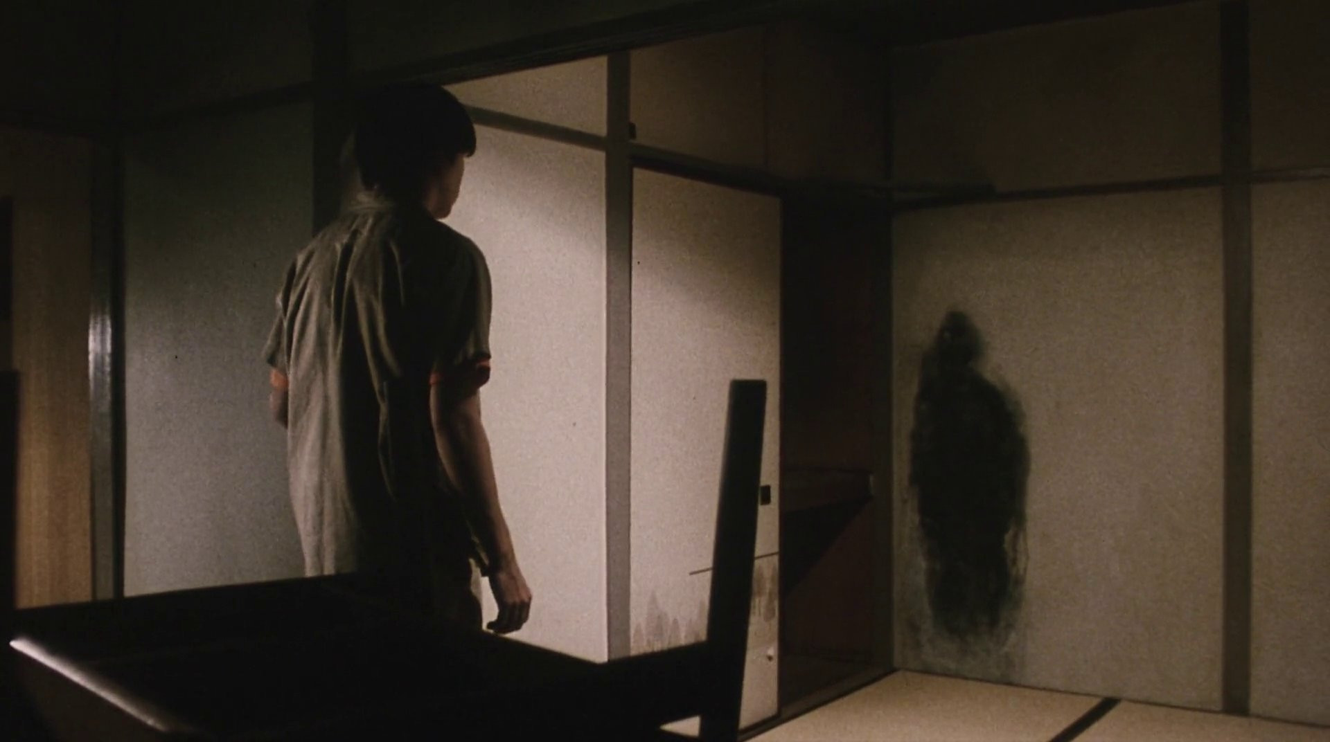 Screenshot from the movie Kairo (2001): a young man has entered a shadowy, muted colored japanese apartment. In the corner of the room, a deep black smudge, vaguely in the shape of a human figure, lurks on the wall. 