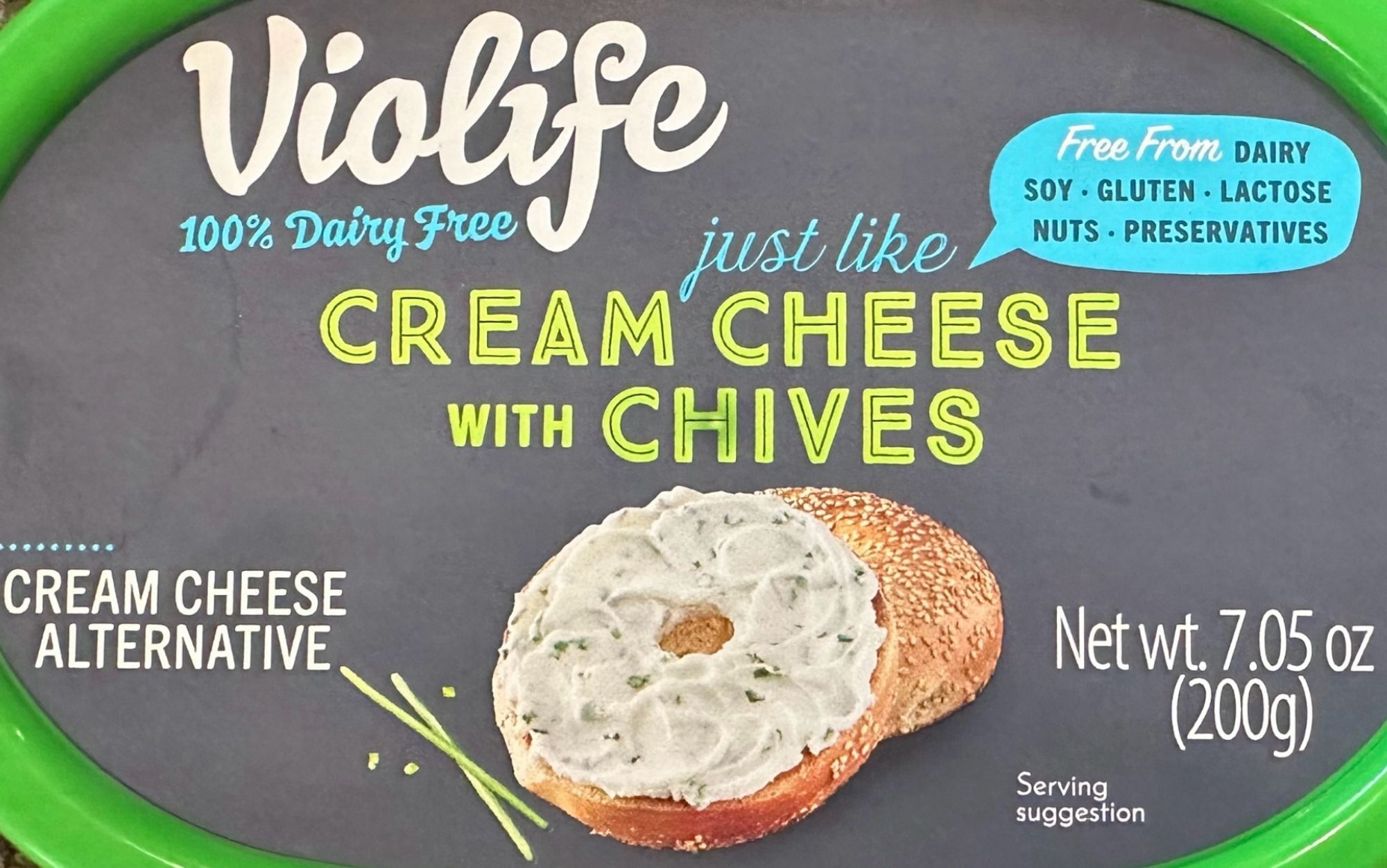 Lid of violife container. Blackboard grey with a green rim. Says Violife 100% Dairy Free just like cream cheese with chives. There’s a little blue speach bubble upper right that says Free from dairy, soy, gluten, lactose, nuts, preservatives. Lower left it says cream cheese alternative. Center bottom is a picture of a bagel with spread on it and some chives lying next to it and the words serving suggestion below & to the right. I will be putting any stray chives ON the bread along with the spread, thank you. Lower right corner it says net weight 7.05 oz (200g). Upon contemplation, the background is probably supposed to remind one more of the slate of a fancy cheeseboard than of a blackboard.