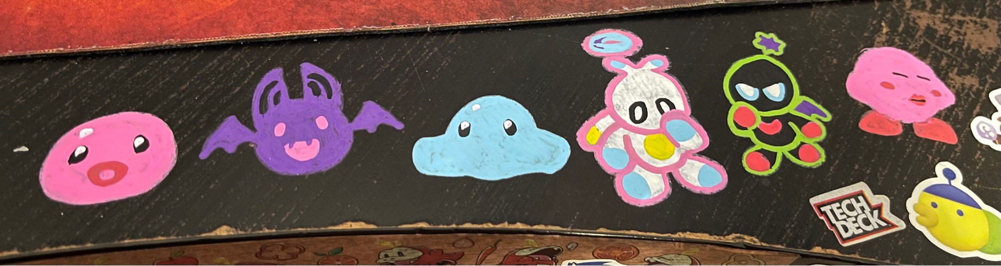 doodles of a Pink Slime, Batty Slime, and Puddle slime from Slime Rancher, as well as a Hero and Dark Chao from Sonic the Hedgehog, and then a weird Kirby, done in Posca pens on a black desk