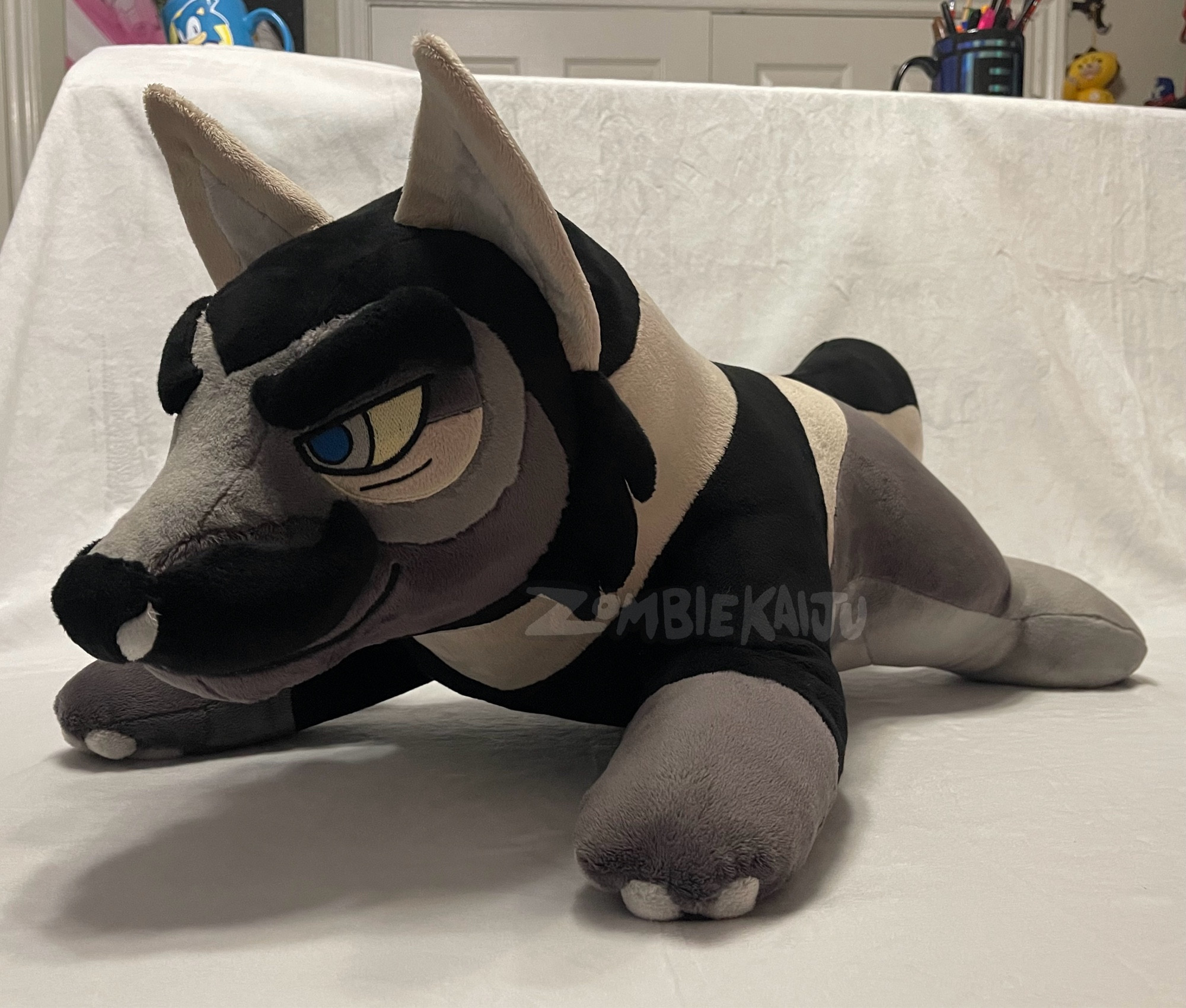 handmade plush of a furry wolf character made of of multiple shades of grey/khaki/black.  Laying on a table facing left side of the screen