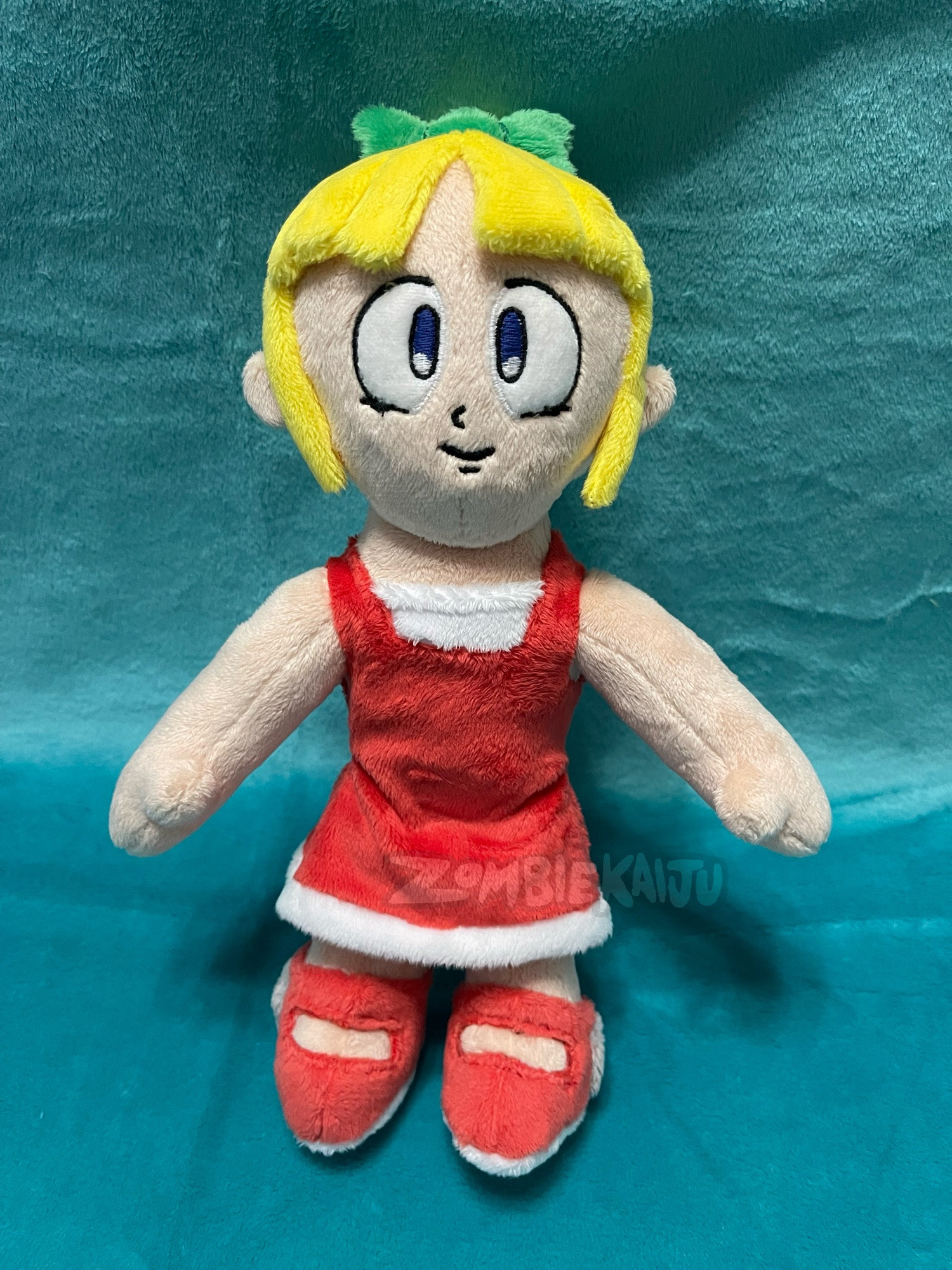 Handmade plush of Roll from Mega Man, a robot girl wearing a red dress, red shoes, with blonde hair and a green hair bow.  Standing in front of a teal backdrop