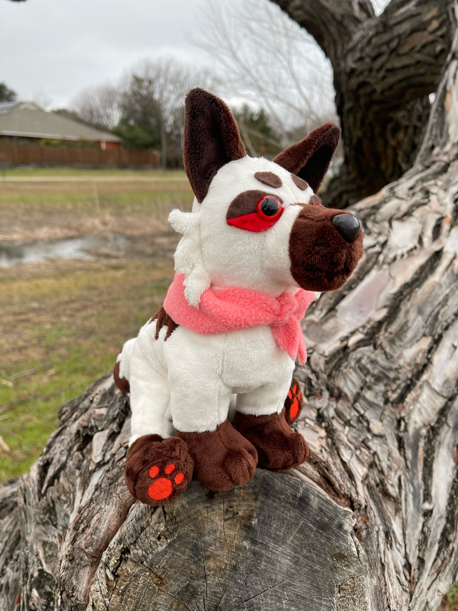 handmade wolf plush cream in color, with brown ears, brown snoot, and brown paws.  paw pads are a bright cherry red, eyes are red with black pupil overtop a brown and red outline.  a coral color bandana is around it’s neck.  plush is in a sitting position, on top of a thick tree branch at a park