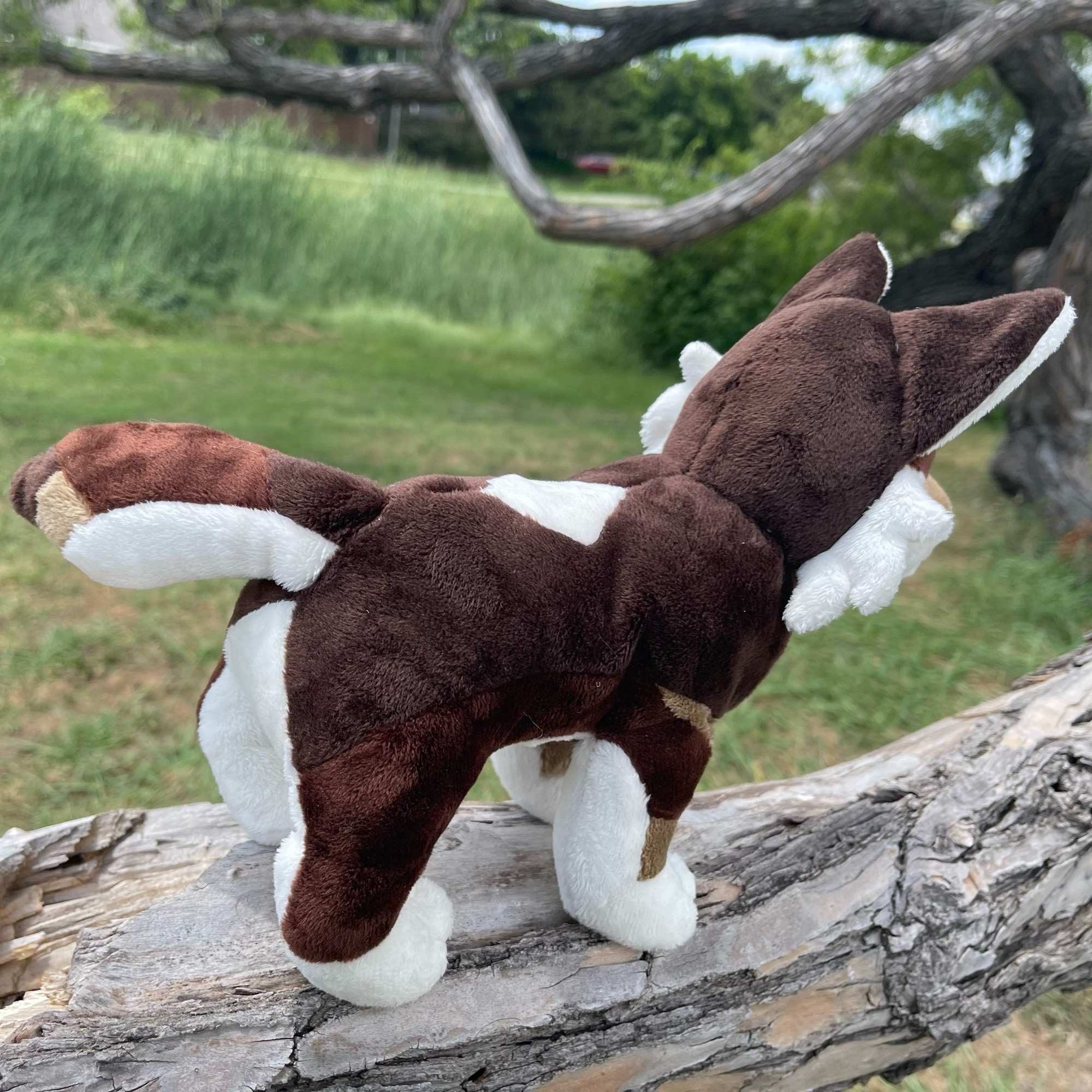 handmade plush of a brown and white wolf, standing on a tree branch, photographed from the side and behind to show the tail