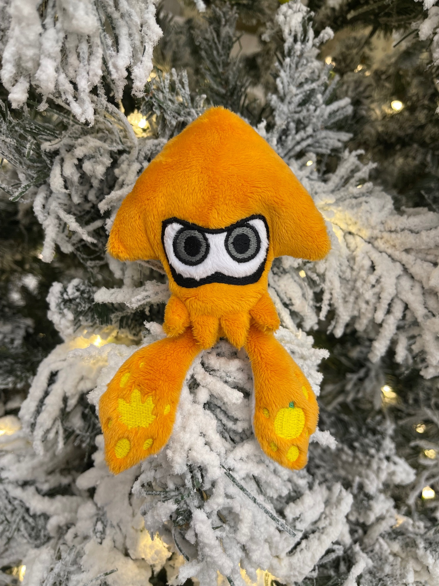 an orange handmade plush of a Splatoon Squid with Fall themed embroidery, sitting on a snow covered christmas tree