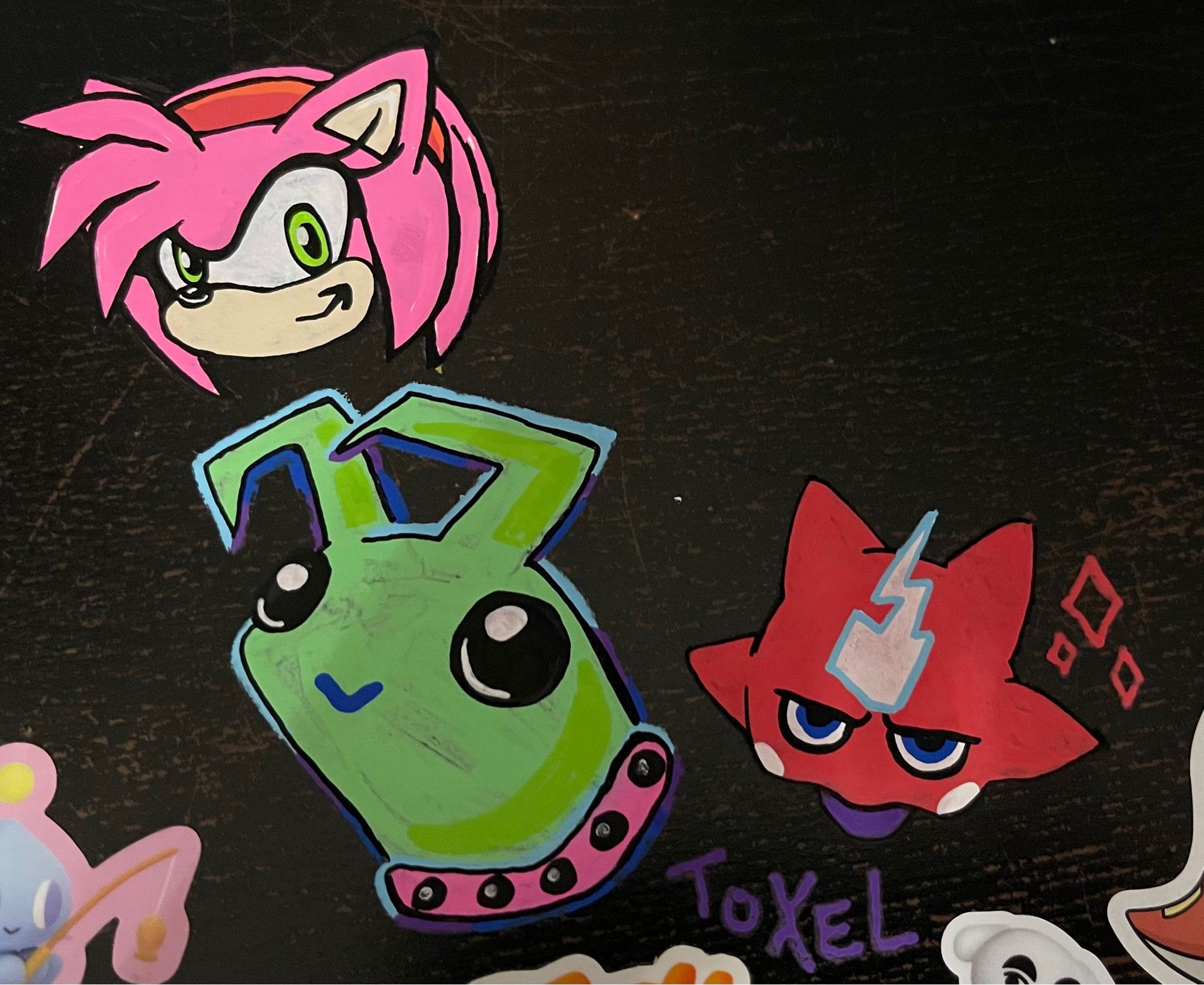 doodles of Amy from Sonic the Hedgehog, a cute green alien creature with a pink collar, and a shiny Toxel from Pokemon, done in Posca pens on a black desk