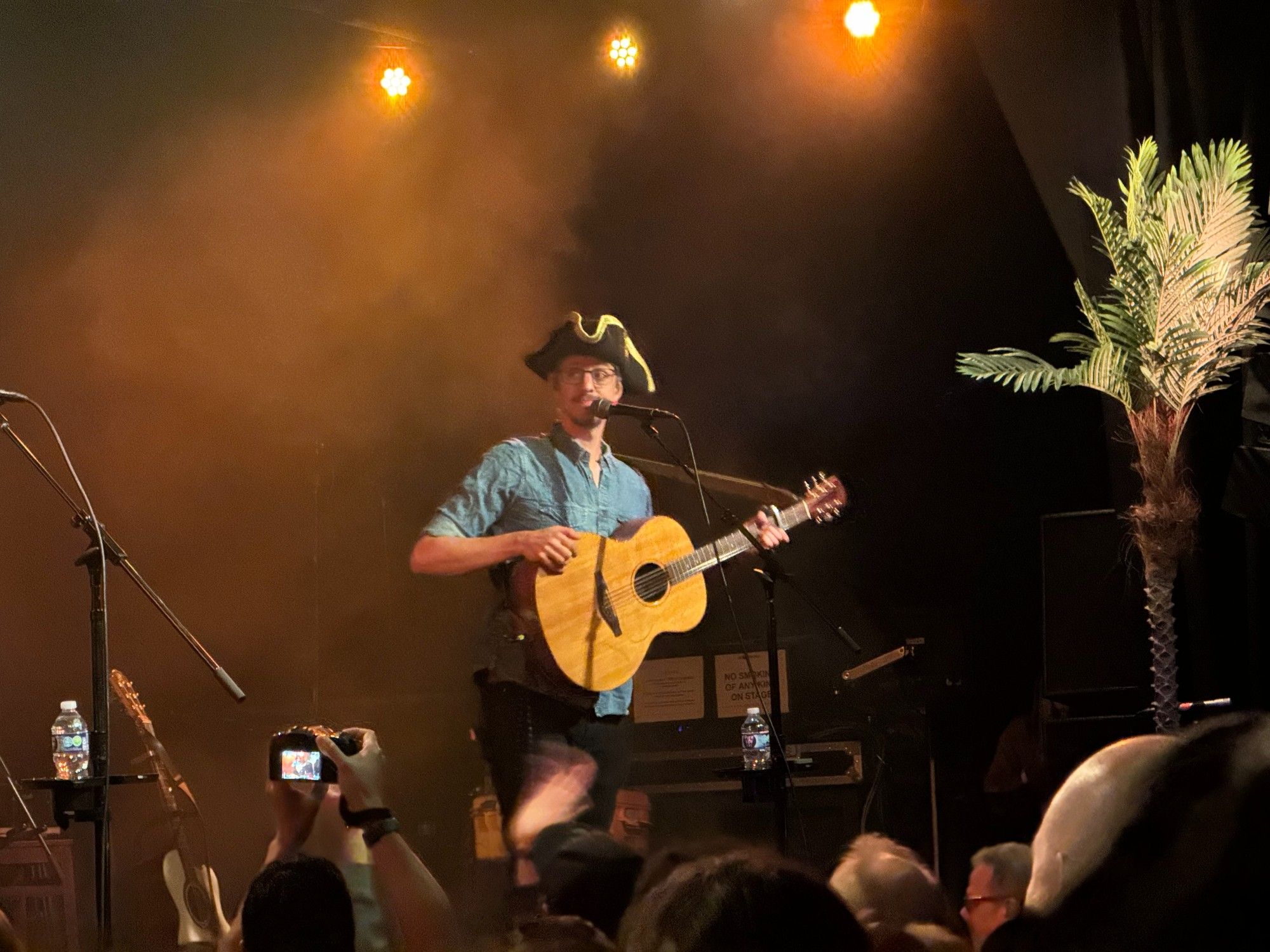 A photo of The Longest Johns' JD Darley, on stage, looking at the rest of the band and wearing a tricorn hat leant to him by an audience member the band decided to refer to as Admiral Beavis McClunkington. A lone, potted palm tree (for morale) is pictured to JD's left.