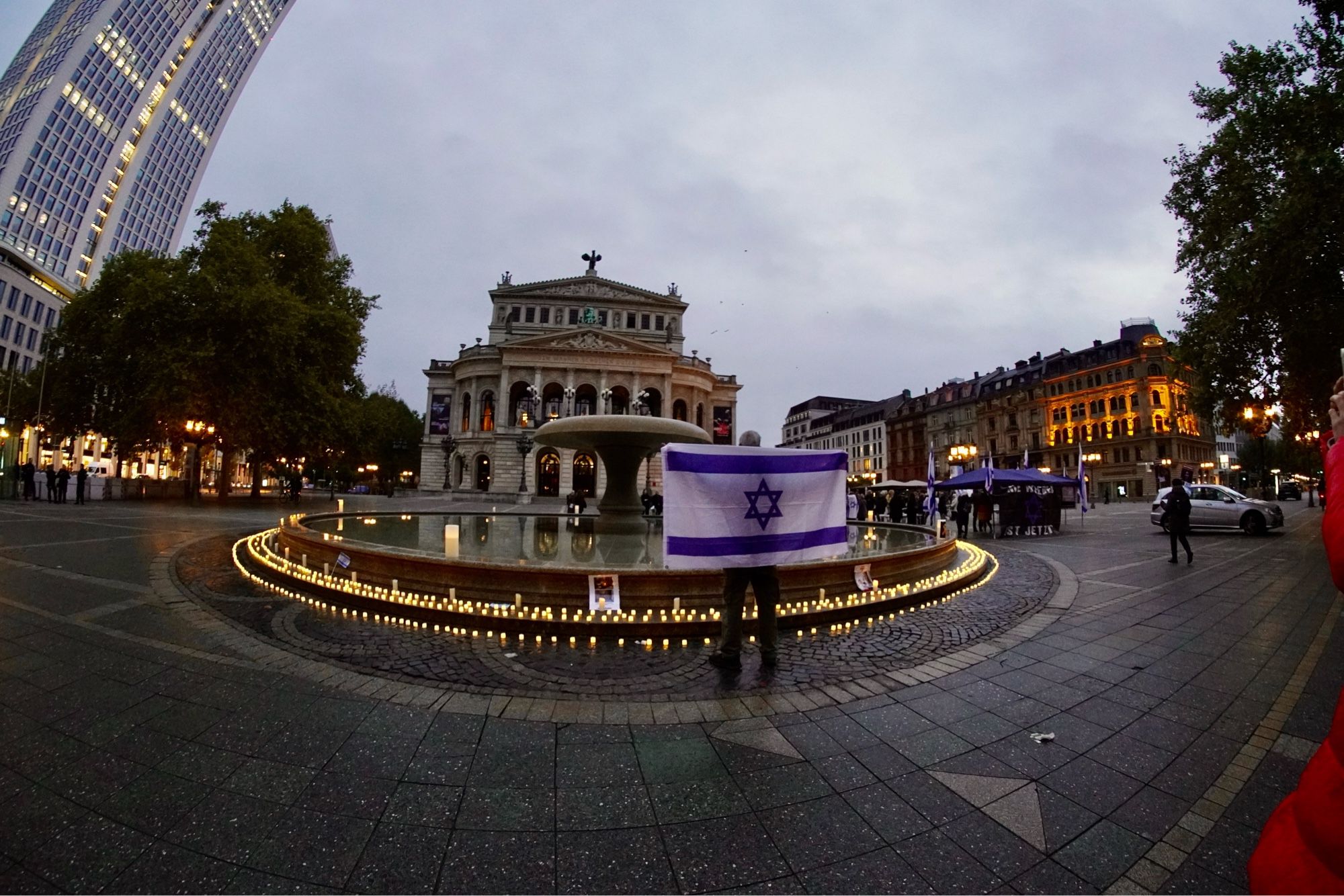 Ein Foto der alten Oper davor ein Mann mit einer israelischen Flagge und einem Kerzenkreis um den Brunnen vor der alten Oper zum Gedenken der Opfer des 7. Oktober.