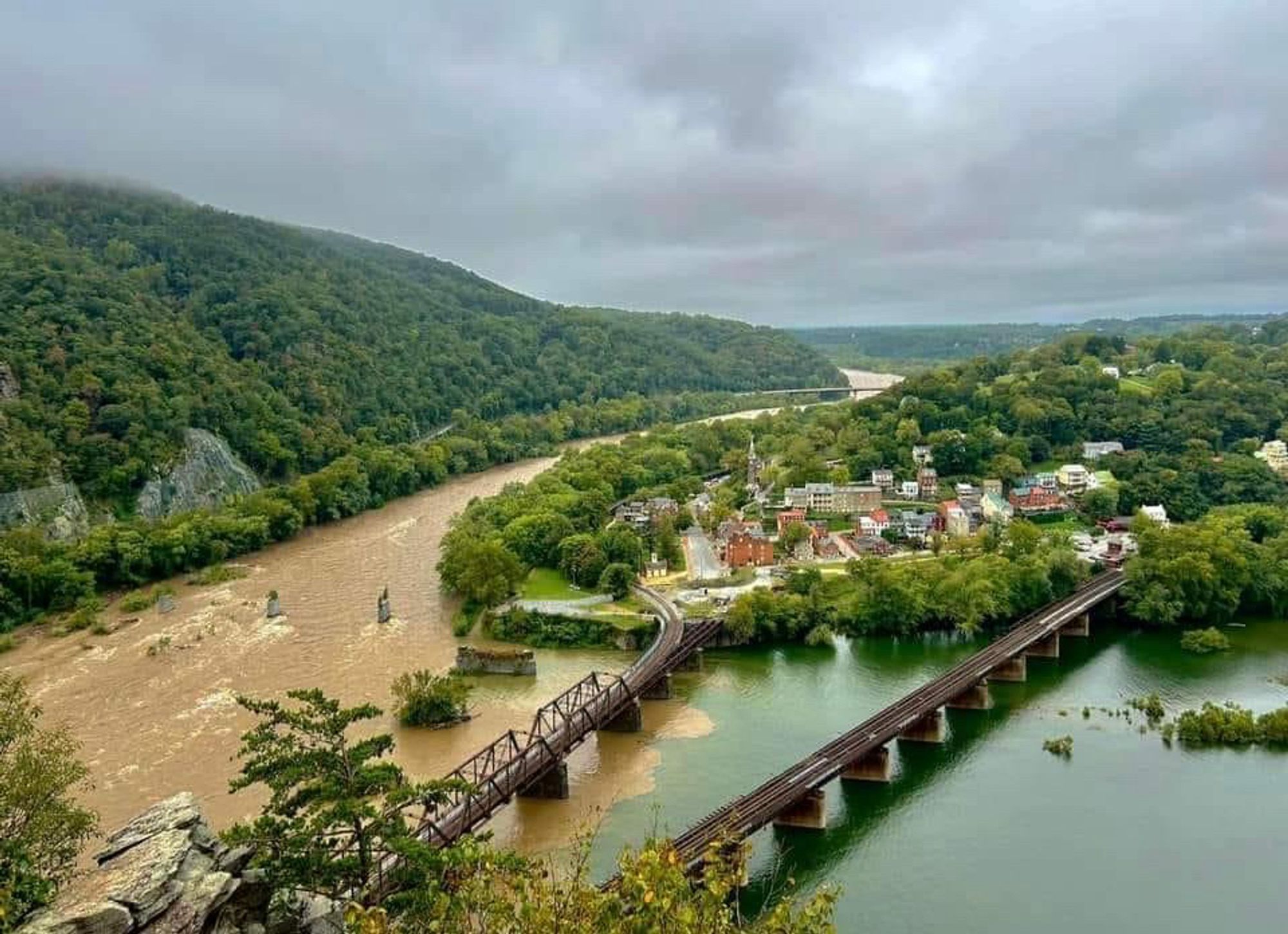 Aerial photo of the Shenandoah and Potomac rivers converging. Green tree-covered hill on the left, the town of Harper’s Ferry in the middle. Two bridges cross the water. The Shenandoah on the left is murky brown; the Potomac is green-blue. A sharp line between them.