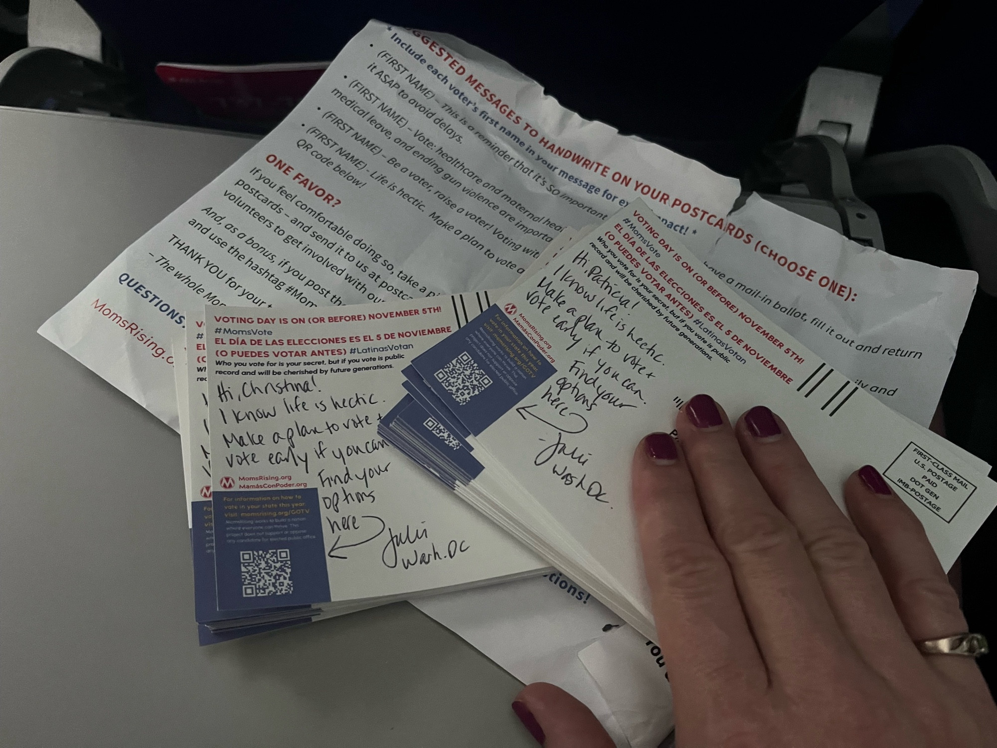 Stacks of postcards with handwritten messages encouraging moms to vote, on an airplane tray table. My hand covers the address of the top card.