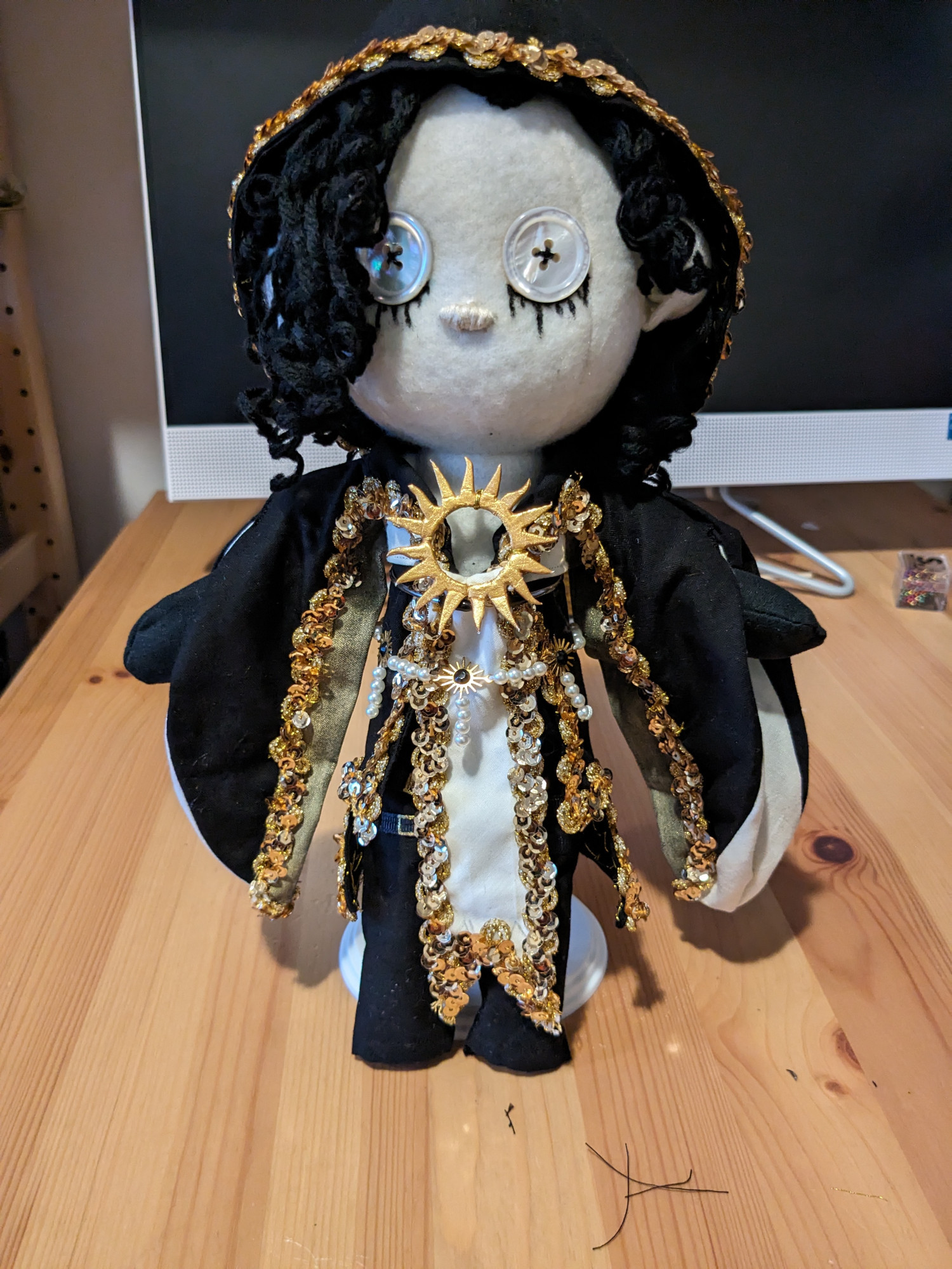 Small pale dollie with black curly hair who looks nothing like me. He wears a thick black hooded robe with gold trim, and a white liner. He has a belt made of pearls and sun beads. The center of his chest has a large sun emblem. He wears black pants with unnecessary but fashion forward belts with gold buckles on said pants. He has pearlescent button eyes and sad boy mascara tear marks underneath them.