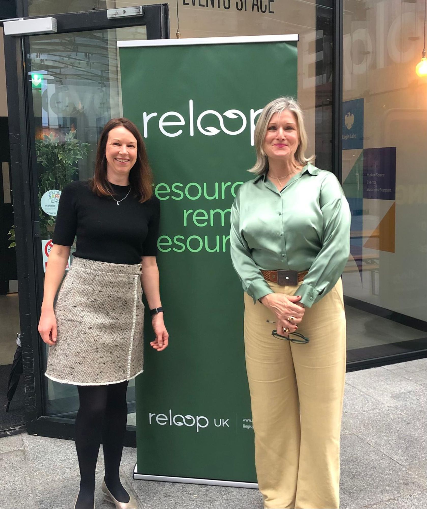 Two women are standing side by side, smiling for the camera in front of a green "Reloop" banner. The woman on the left is wearing a black top and a tweed skirt, while the woman on the right is wearing a light green satin blouse and beige trousers.