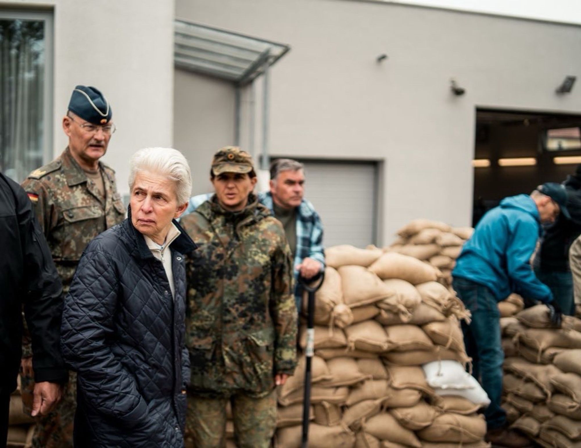 Marie-Agnes Strack-Zimmermann macht sich im Beisein von Soldaten einen Eindruck von der Lage in den Hochwassergebieten. Im Hintergrund sieht man Sandsäcke und ehrenamtliche Helfer.