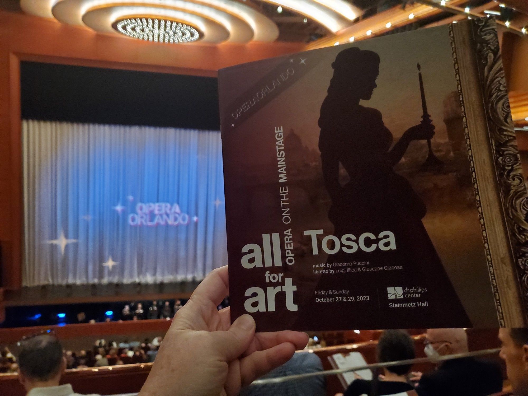 Photo of opera program Tosca, held up to background of Orlando Opera sign at Steinmetz Hall