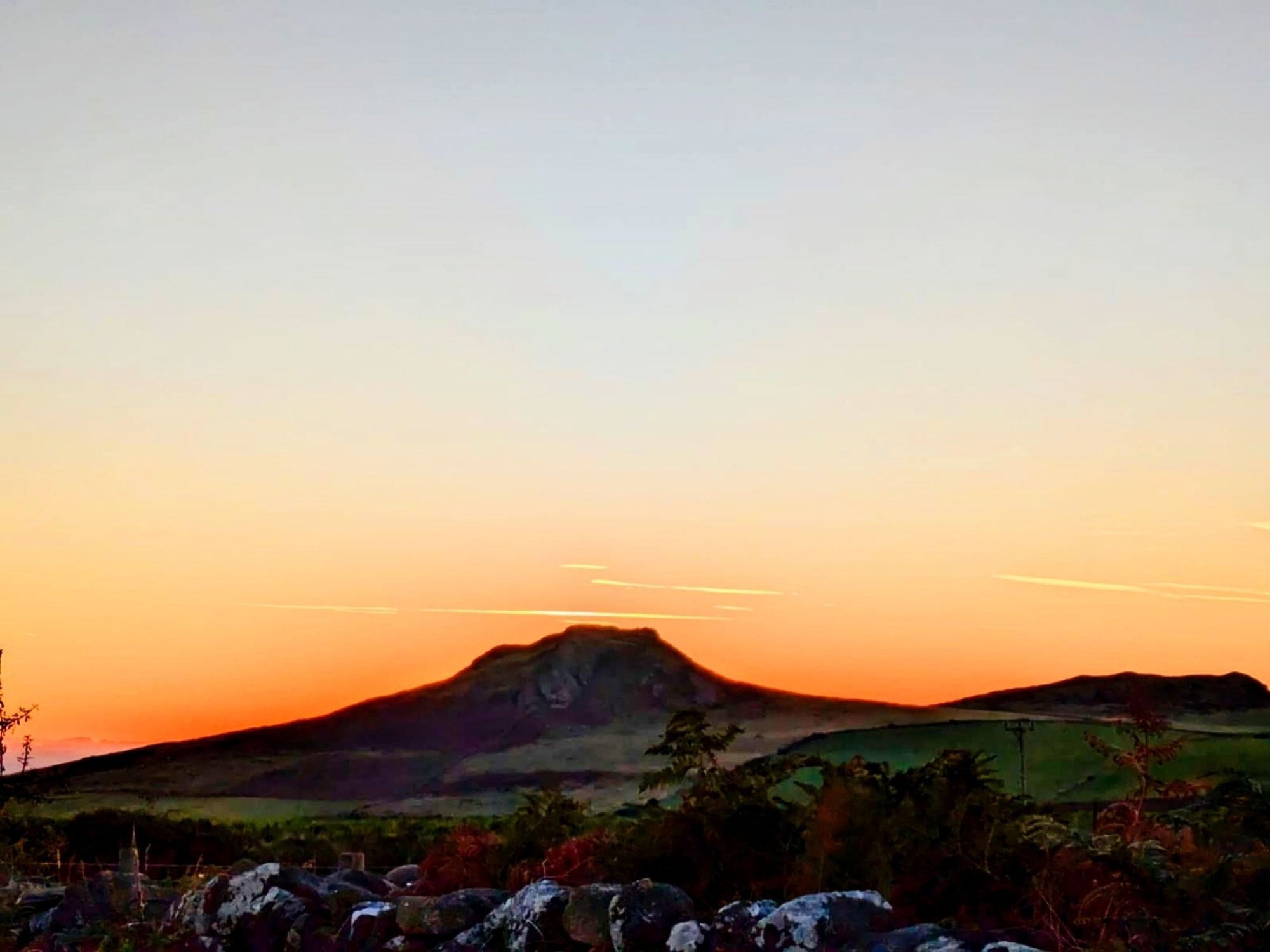 Sun rising but obscured by a silhouette of a hill. Fields are patchwork green and there is a stone wall in the foreground. Background sky graduates from orange to very light blue.